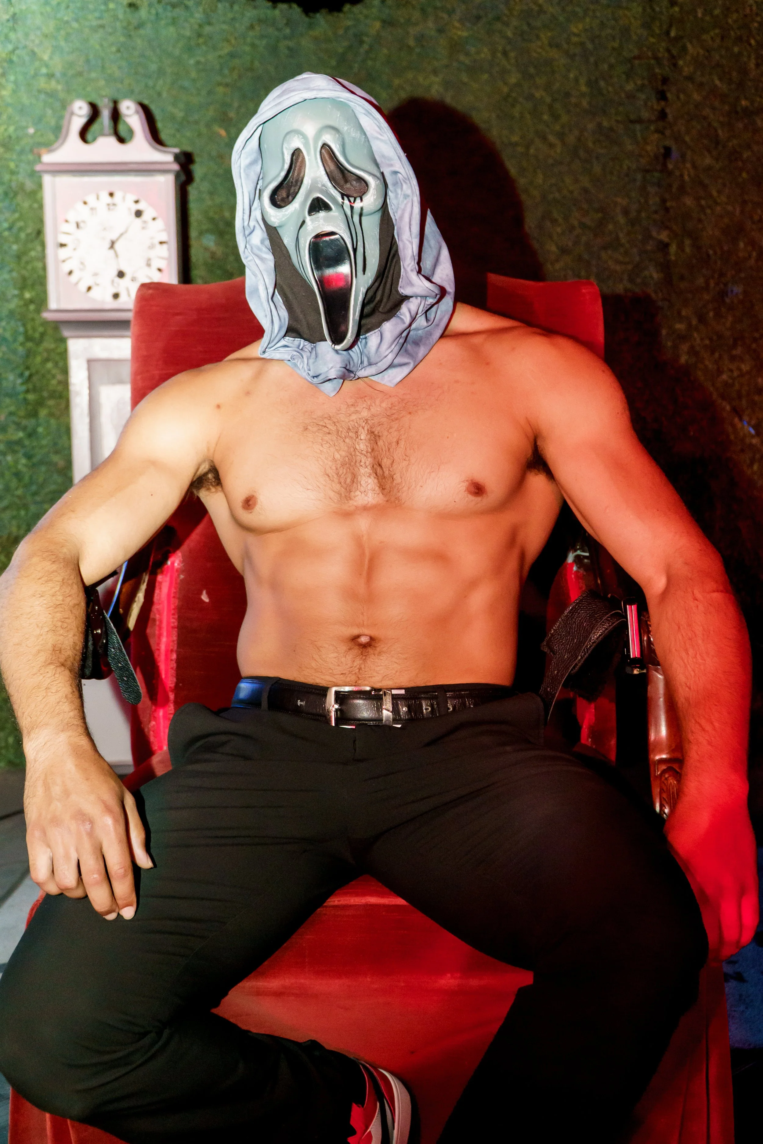 A shirtless man wearing a Ghostface mask from the movie Scream, sitting on a red armchair with a green background and a clock in the background.