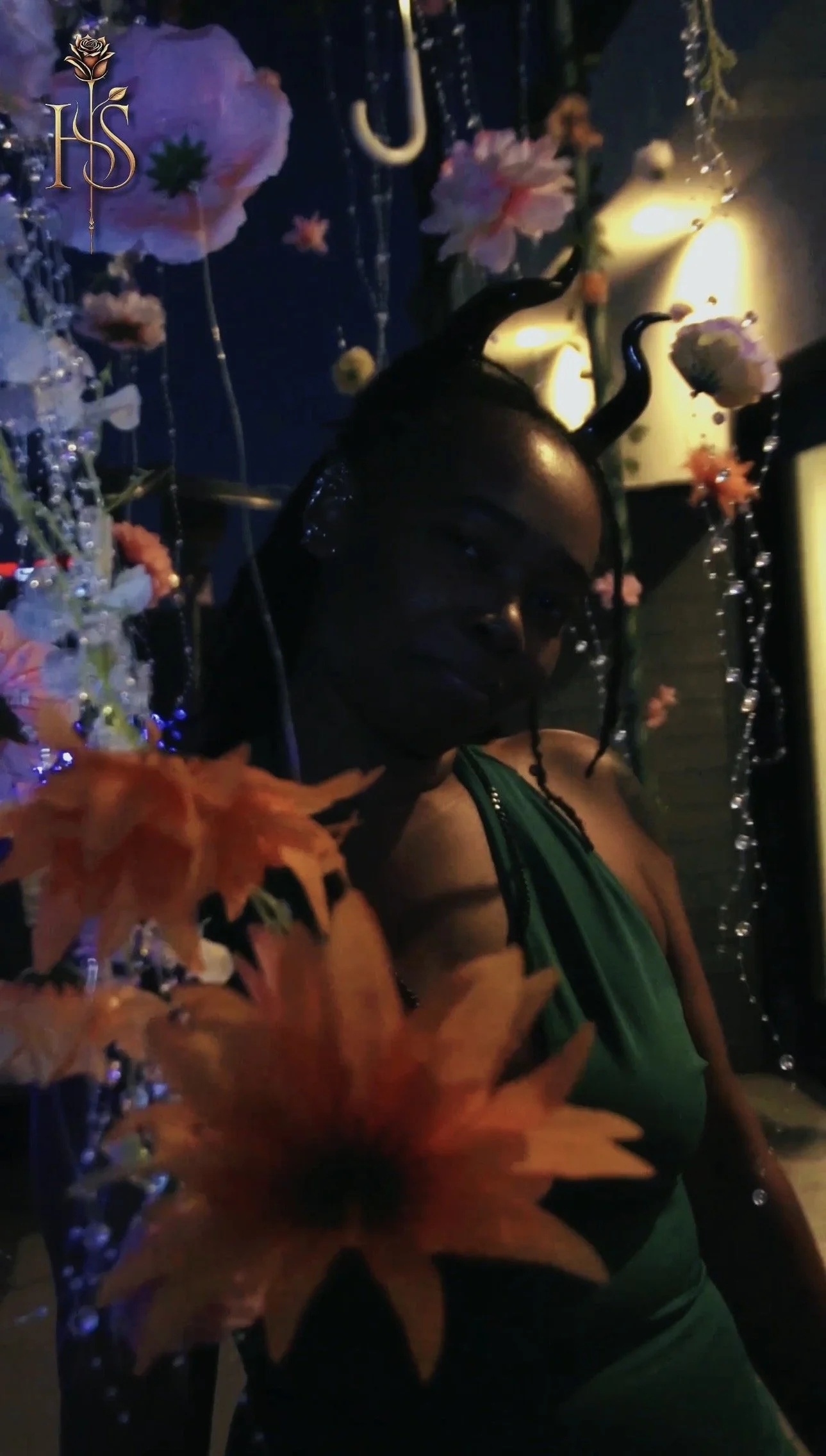 A woman with dark hair in a green dress posing at a decorated event with flowers, beads, and string lights, in low lighting.