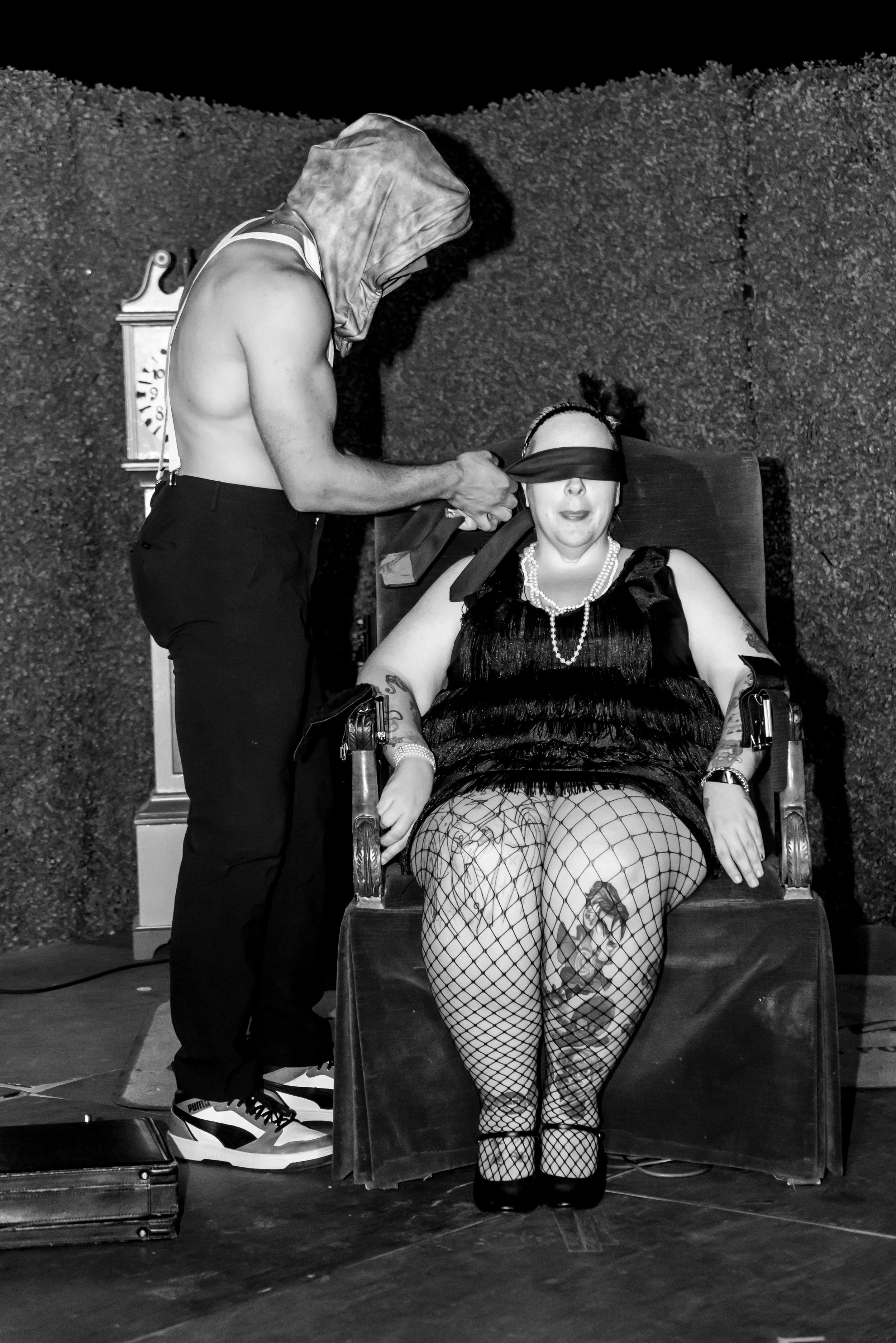 A person with a hood and no shirt, standing next to a woman seated in a chair. The seated woman is blindfolded, wearing a dress, pearl necklaces, fishnet stockings, and has tattoos. The setting appears to be an outdoor or stage area with a textured w