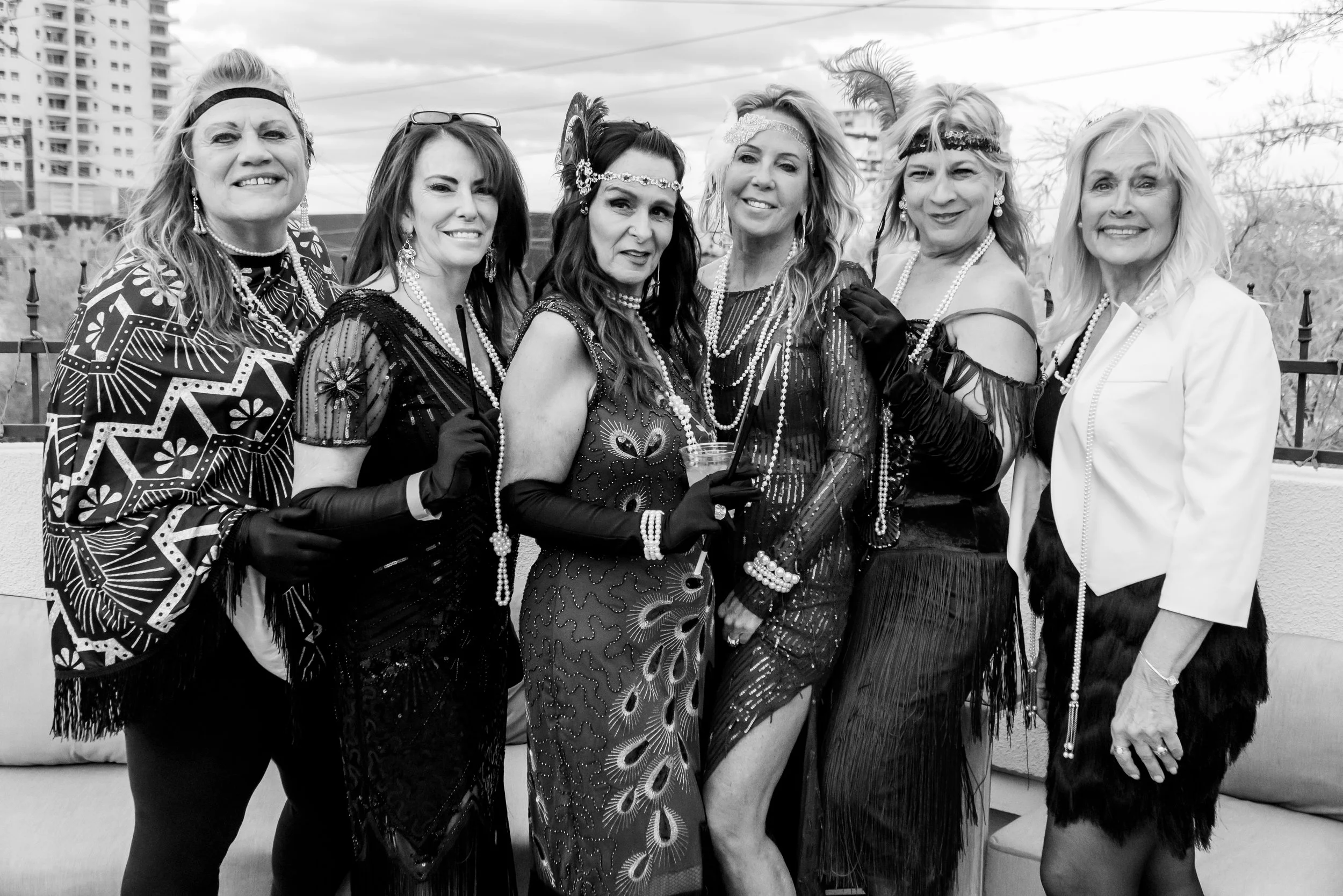 Group of seven women dressed in flamboyant, 1920s-style costumes with feathers, beads, and accessories, posing at an outdoor event in an urban area with buildings visible in the background.