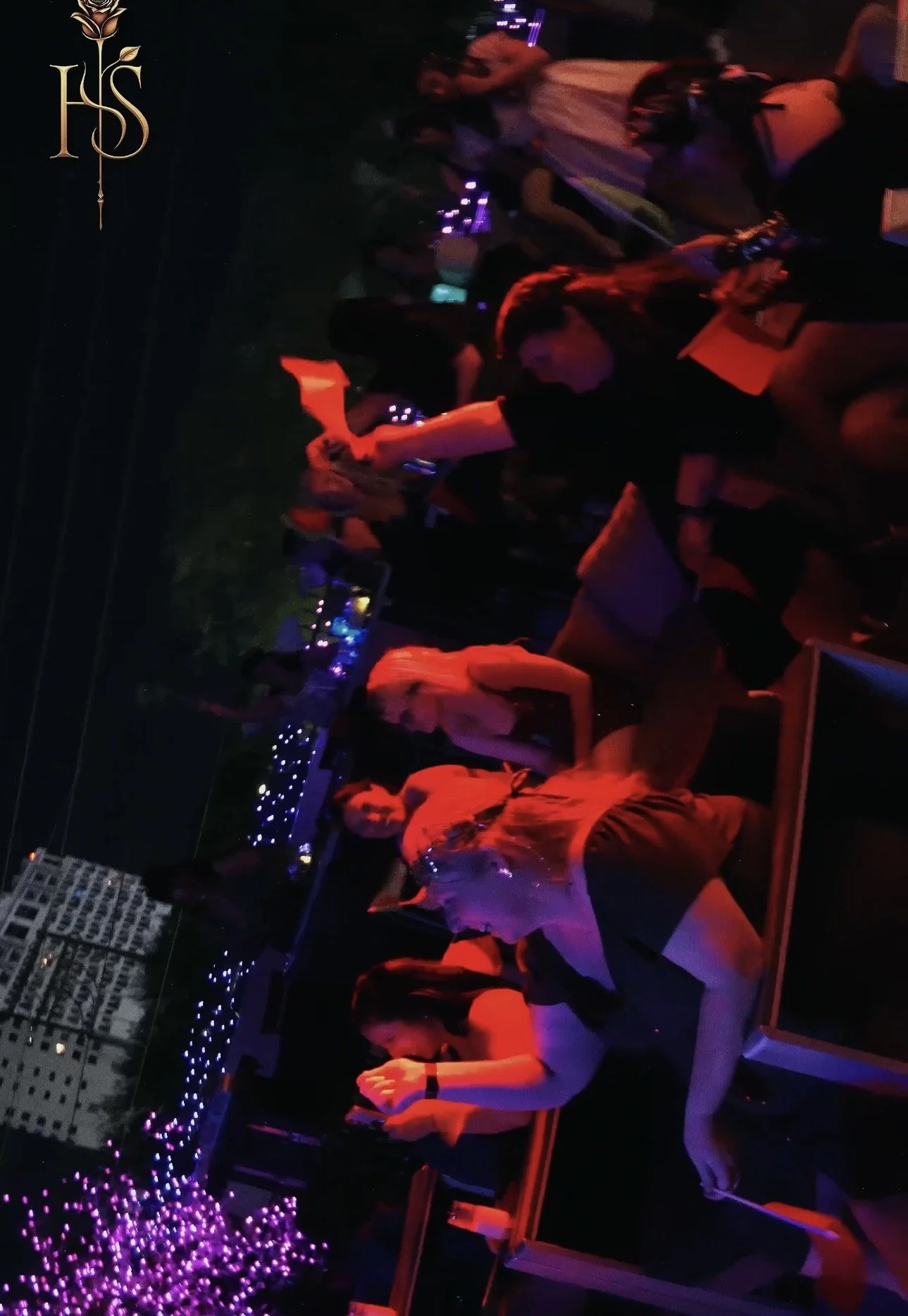 People sitting on a balcony at night, illuminated by purple and red lights, with city buildings in the background.