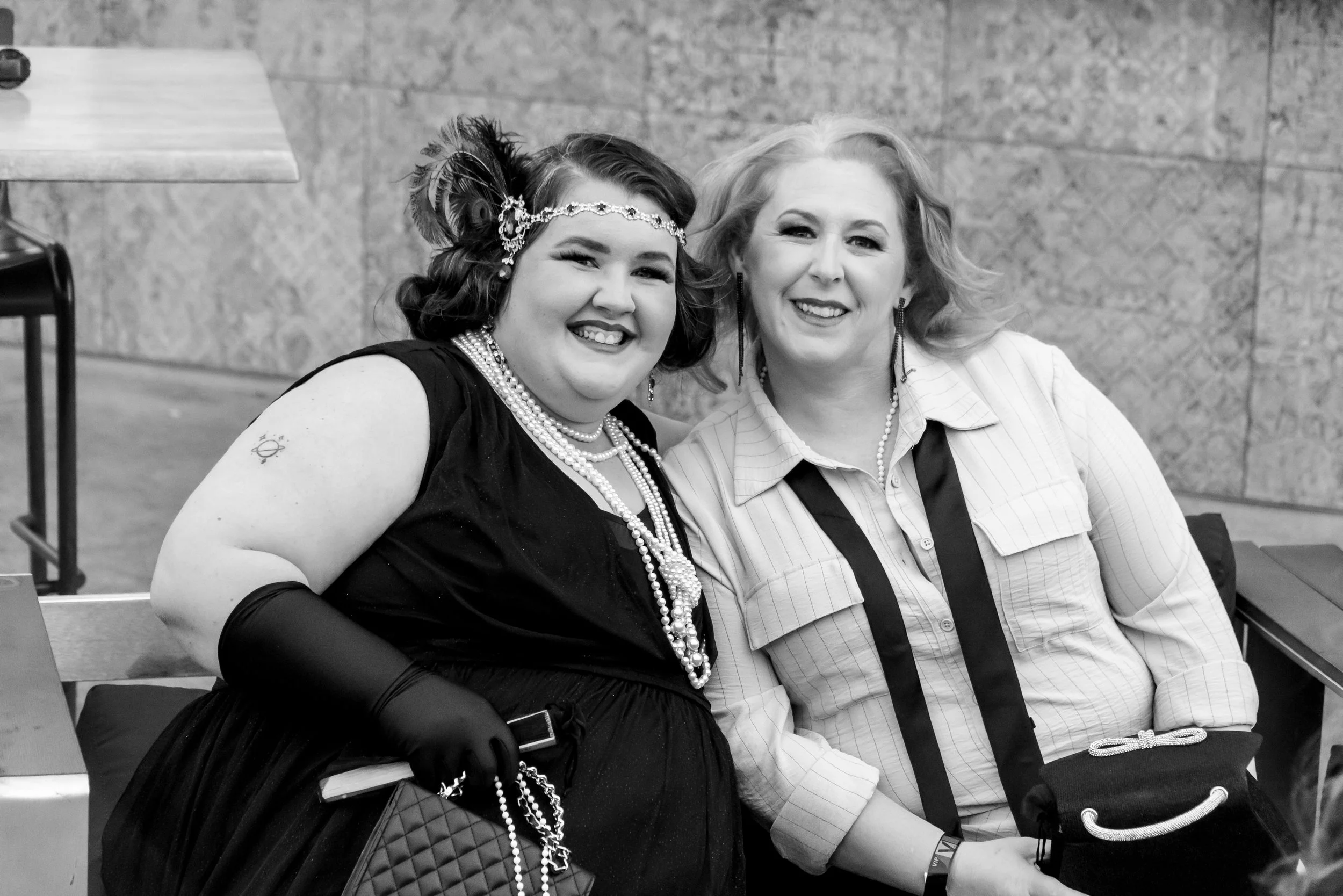 Two women sitting close together, smiling for the camera. One woman has short light-colored hair and is wearing earrings, a striped shirt, and a black tie. The other woman has dark hair styled in vintage waves, adorned with a feathered headpiece and 