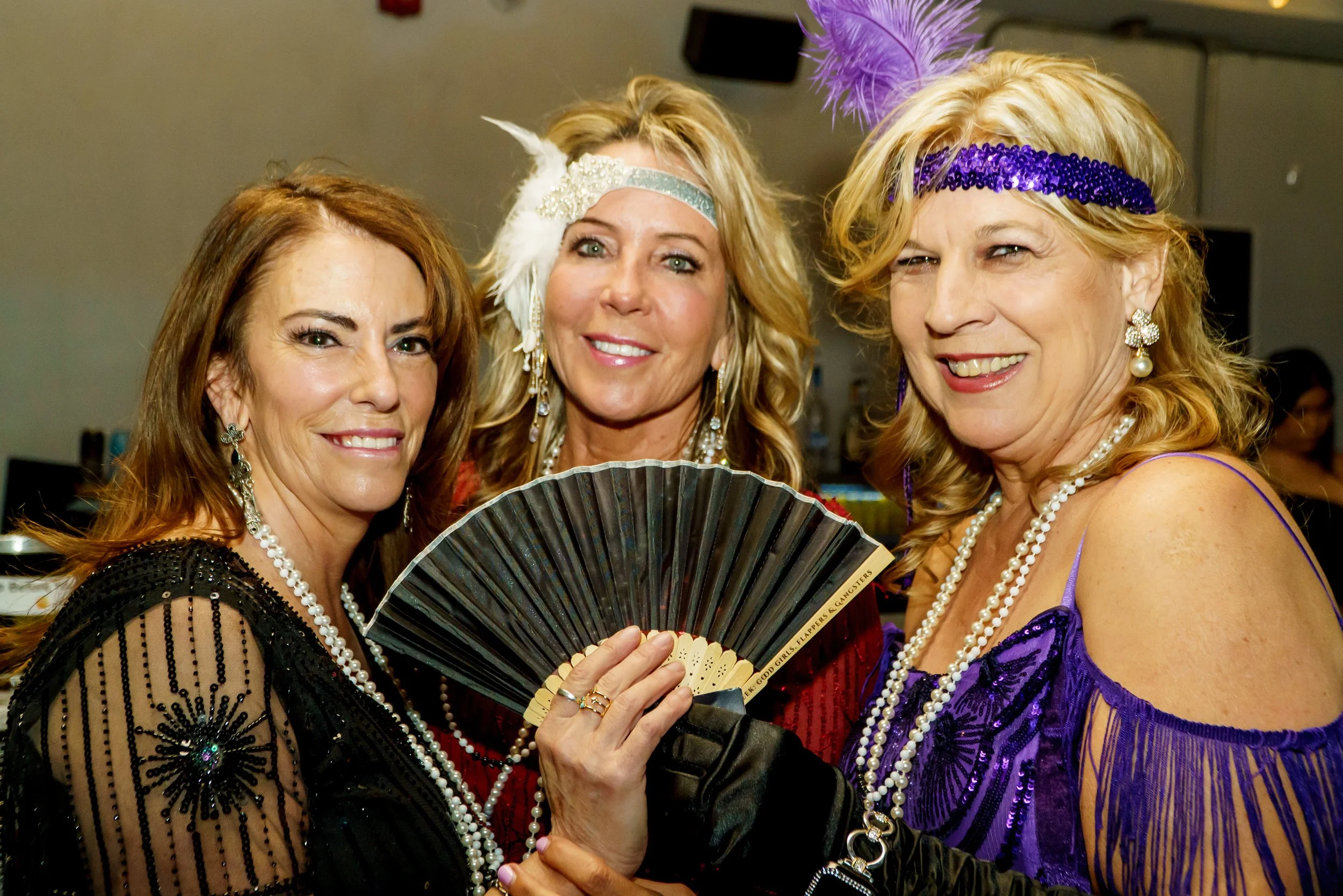 Three women dressed in colorful, sparkly costumes with pearl jewelry, holding a decorative fan, smiling for the camera at a festive event.