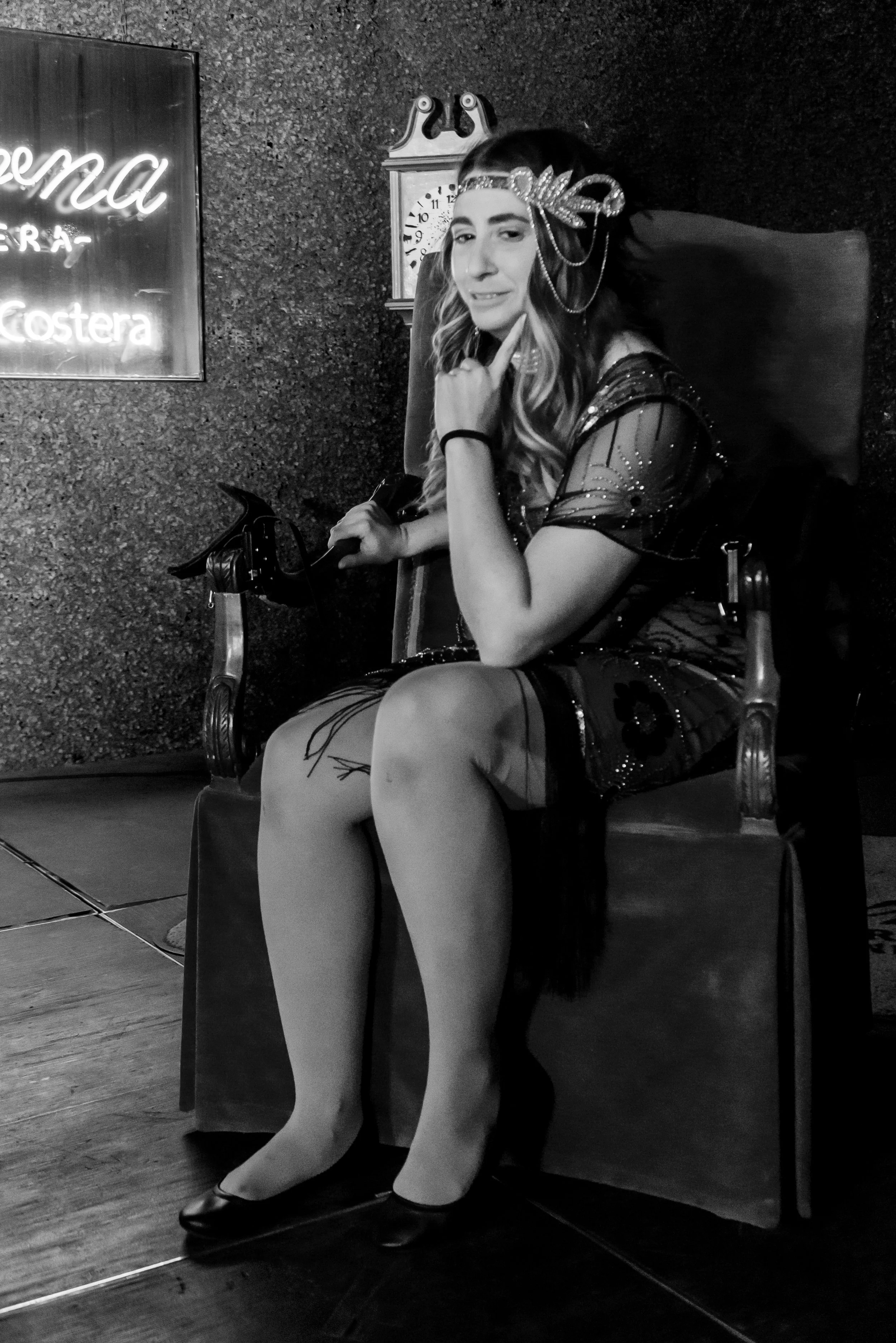 A woman sitting on a chair at a restaurant, touching her chin and looking at the camera, wearing a decorative headpiece and a stylish dress. There is a wall clock and a neon sign on the wall behind her.