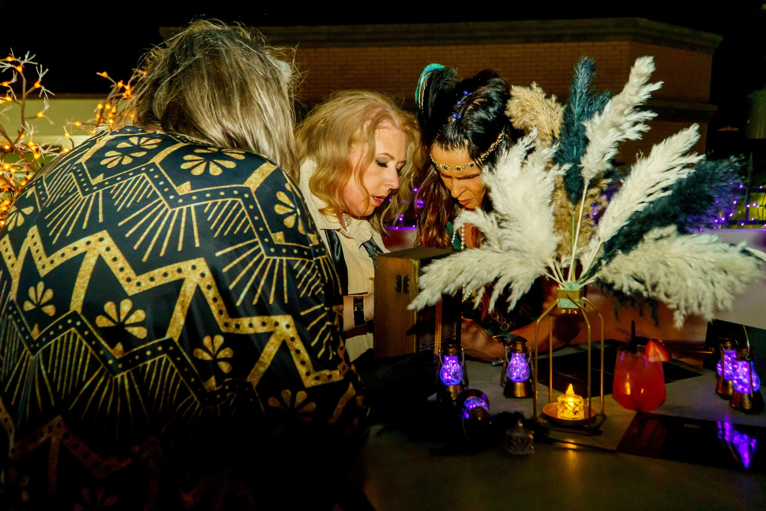 Three women looking at a display with feathers and lamps, decorated with string lights at night.