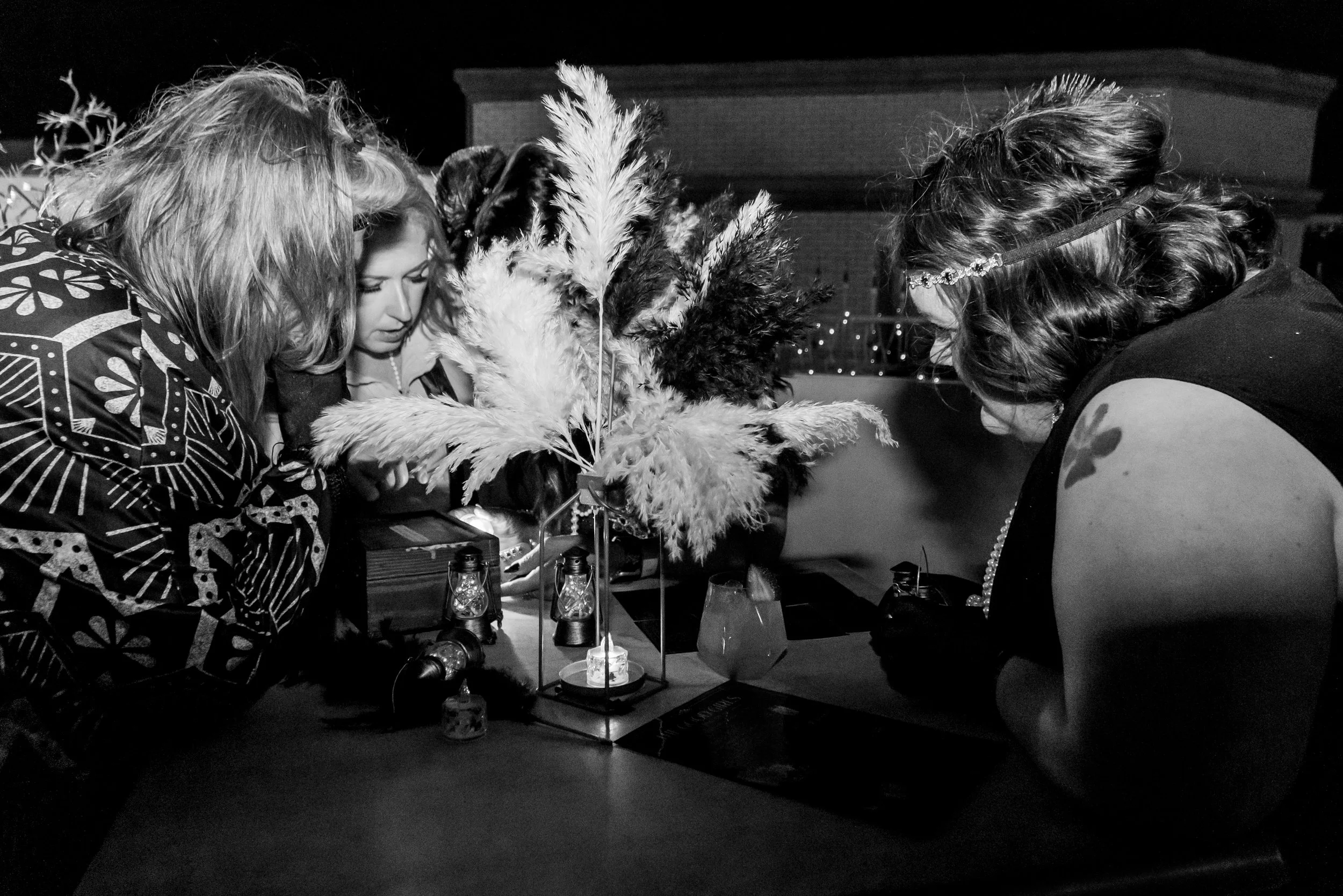 Three women gathered around a table with feathers and lanterns, engaged in an activity, possibly craft or ritual, outdoors at night.