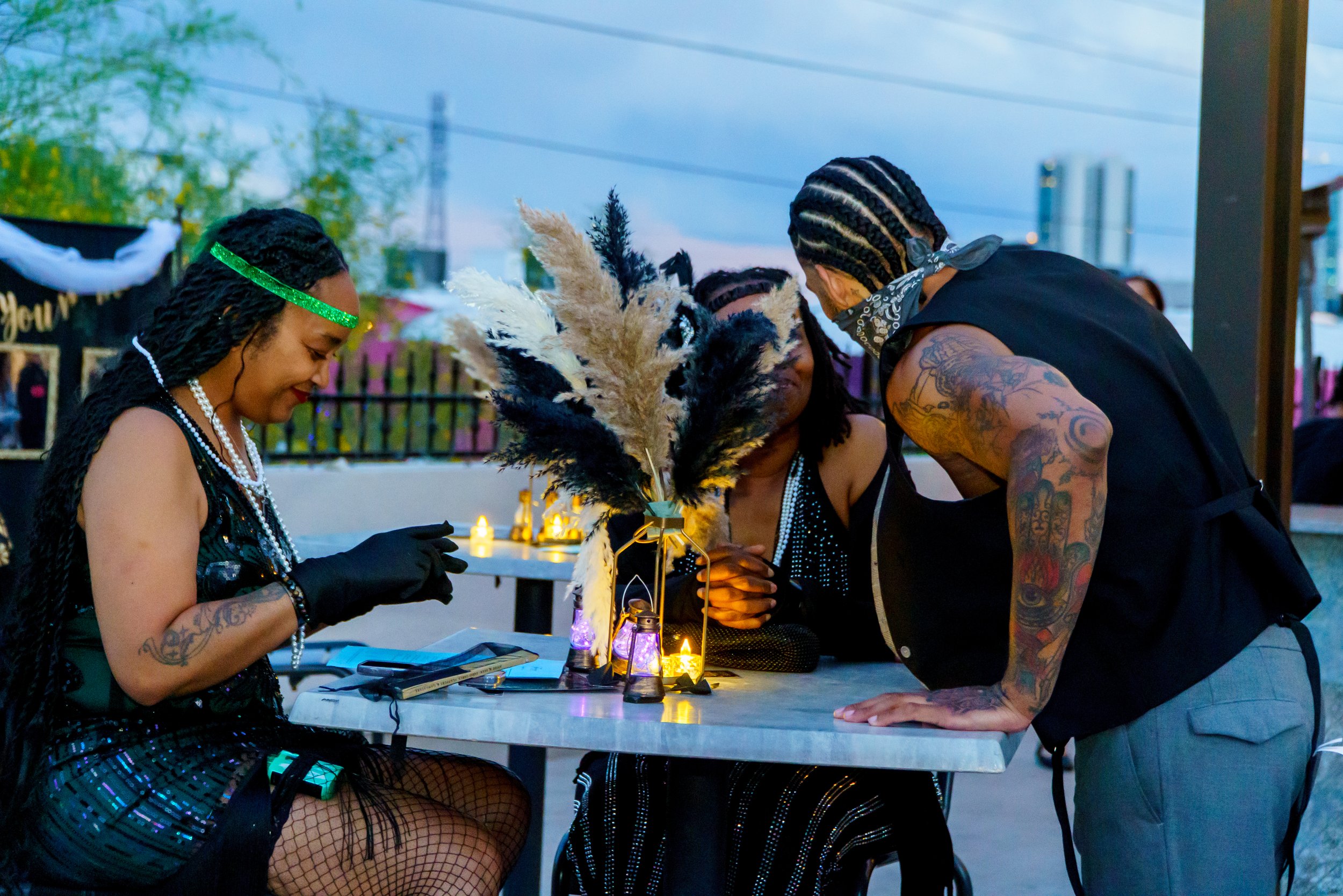 Three people with tattoos and dark clothing gather around a table with lit candles, feathers, and decorative items during a dusk outdoor event.