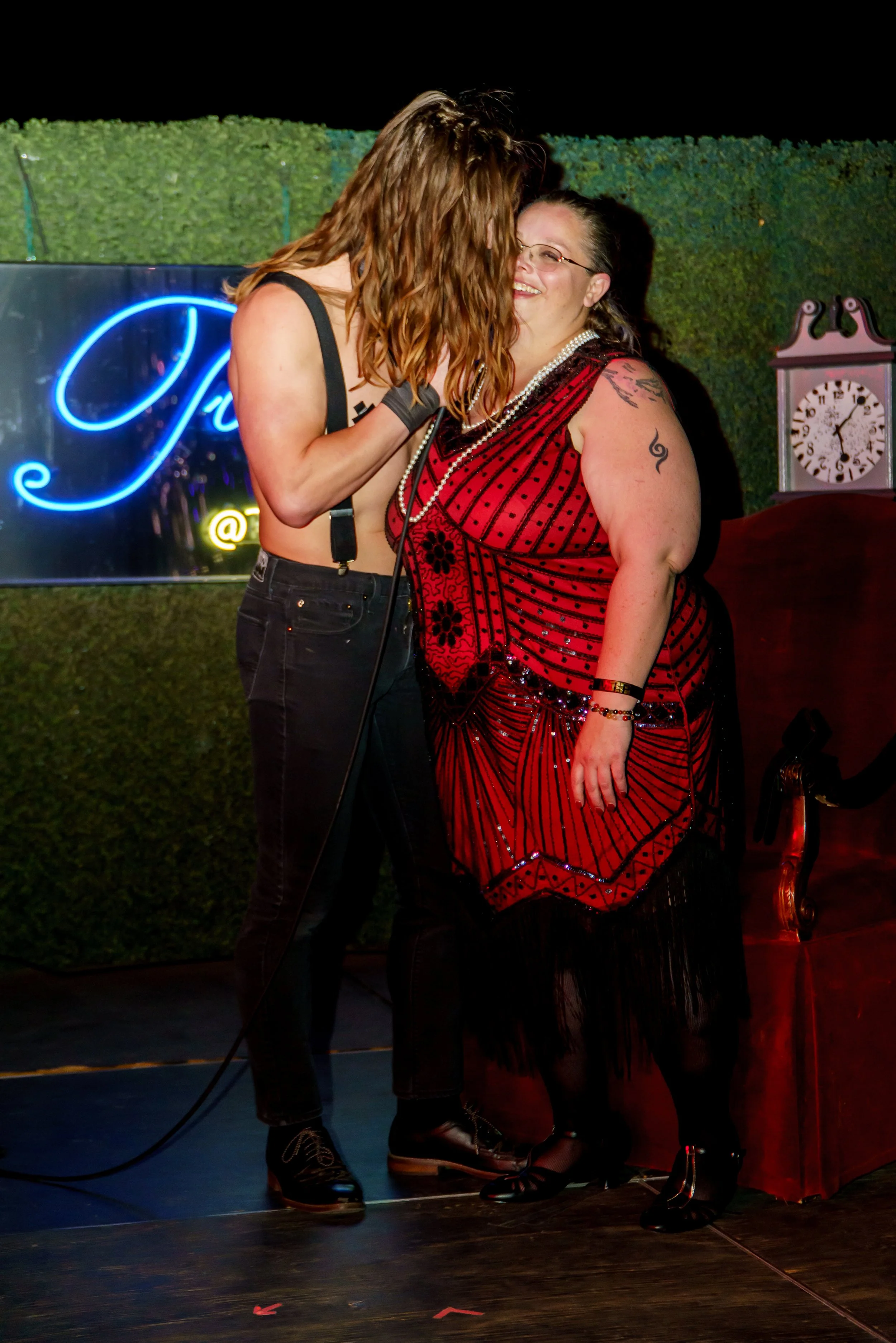 Two women sharing a joyful moment at a social event, with one woman wearing a red dress with black designs and the other in a black crop top, in front of a blue neon sign and a decorative clock.