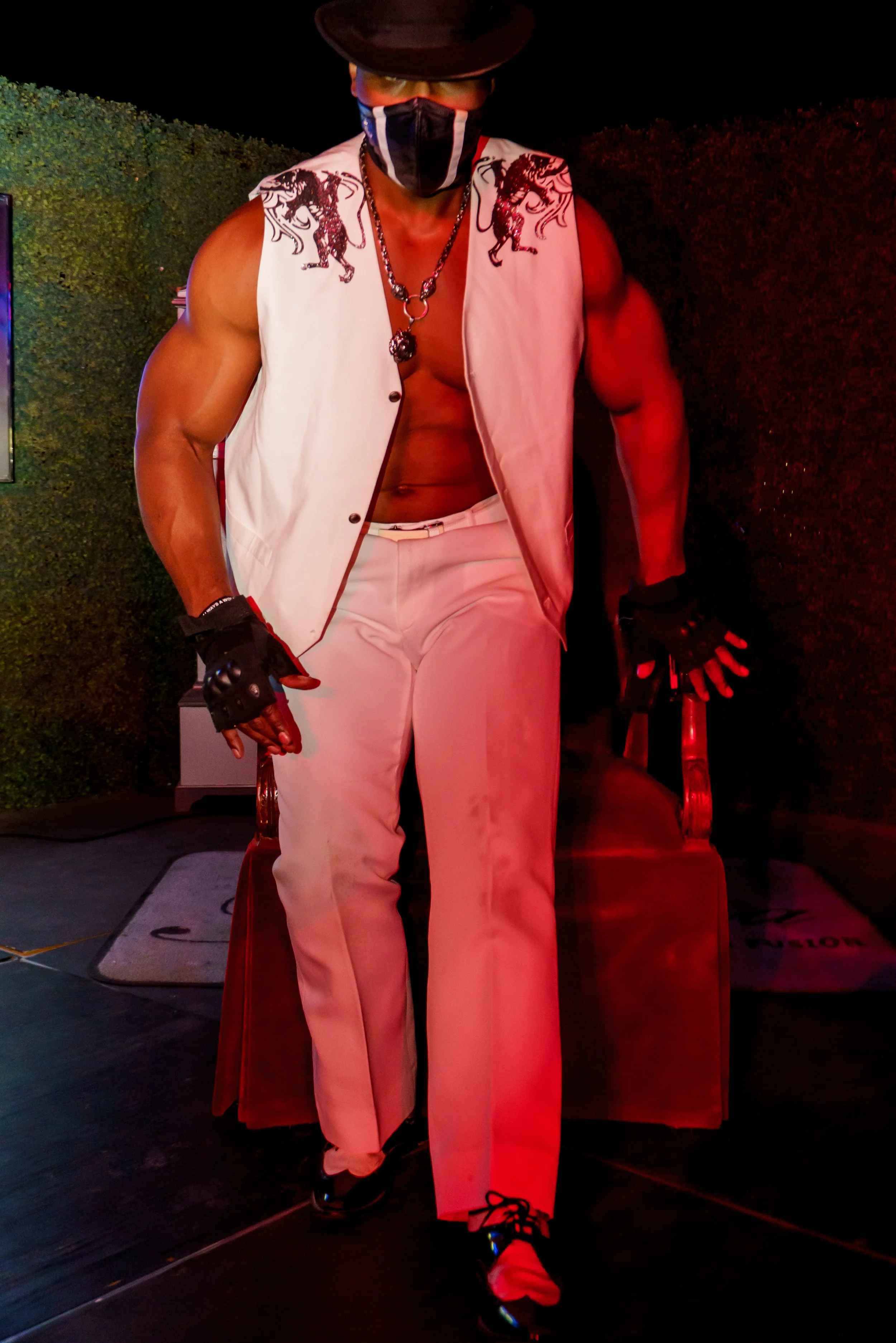 A man dressed in white pants and a sleeveless jacket with lion embroidery on the shoulders, wearing a black hat, black gloves, a face mask, and a necklace, standing in a dimly lit space with red lighting.