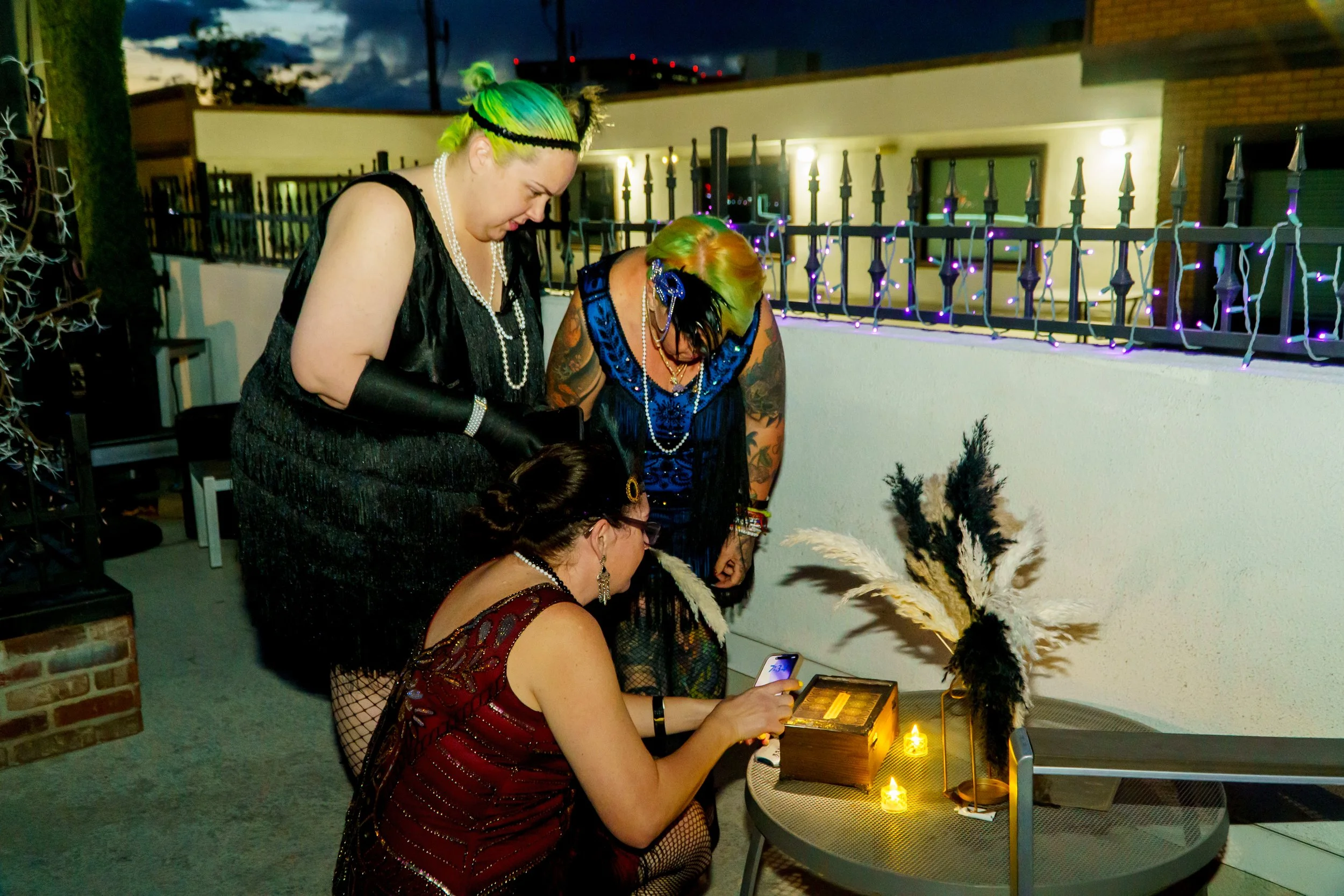 Group of people dressed in vintage attire at an outdoor evening party, with decorative lights, a small table with candles, and festive feather decorations.