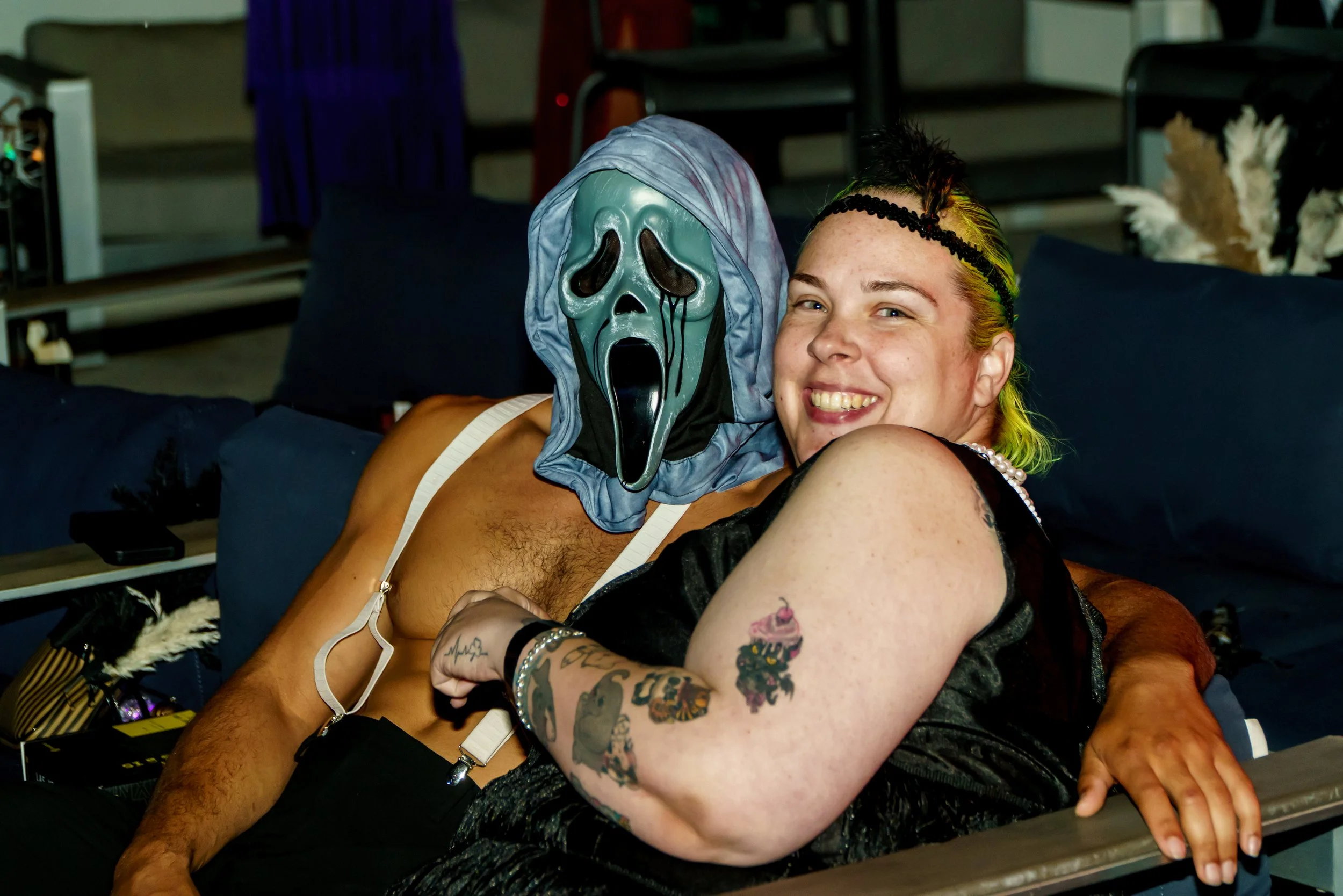 Two people sitting closely together, smiling at the camera. One is wearing a Jason Voorhees mask from the Scream movie series, and the other has colorful, rainbow-like hair with tattoos on her arms.