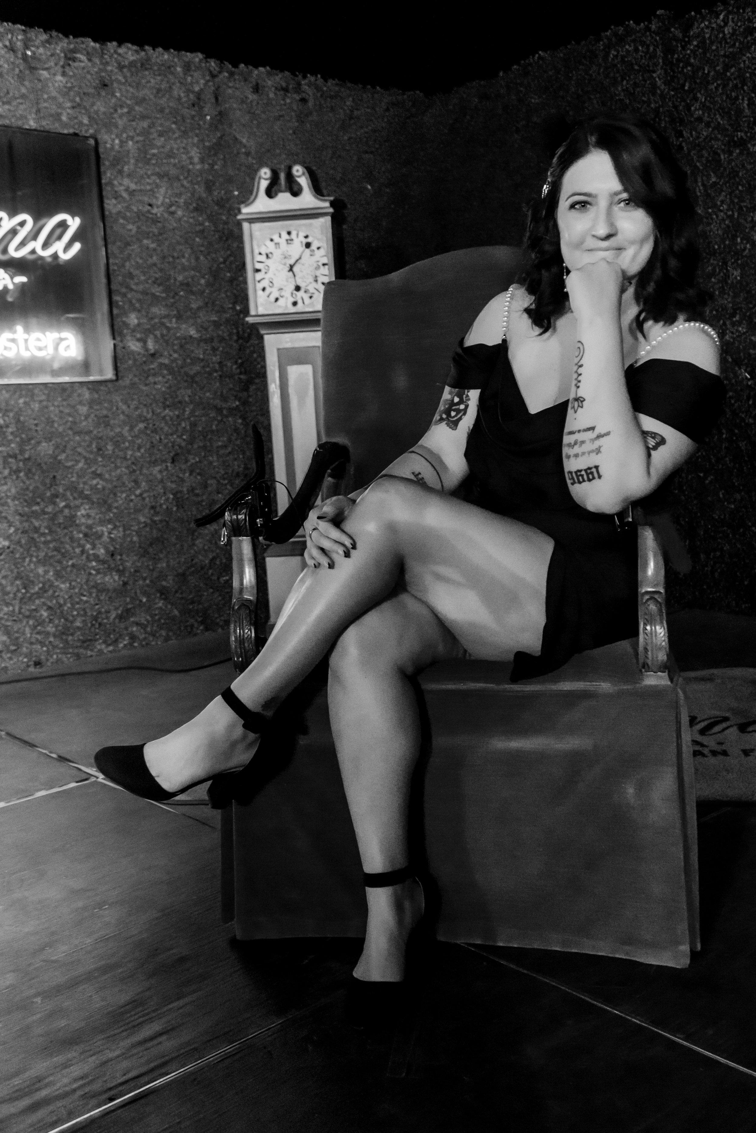 Black and white photo of a woman with dark wavy hair sitting on a chair indoors. She is wearing a short black dress with pearl straps, high heels, and has tattoos on her arms. She is resting her chin on her hand and smiling slightly at the camera. Be