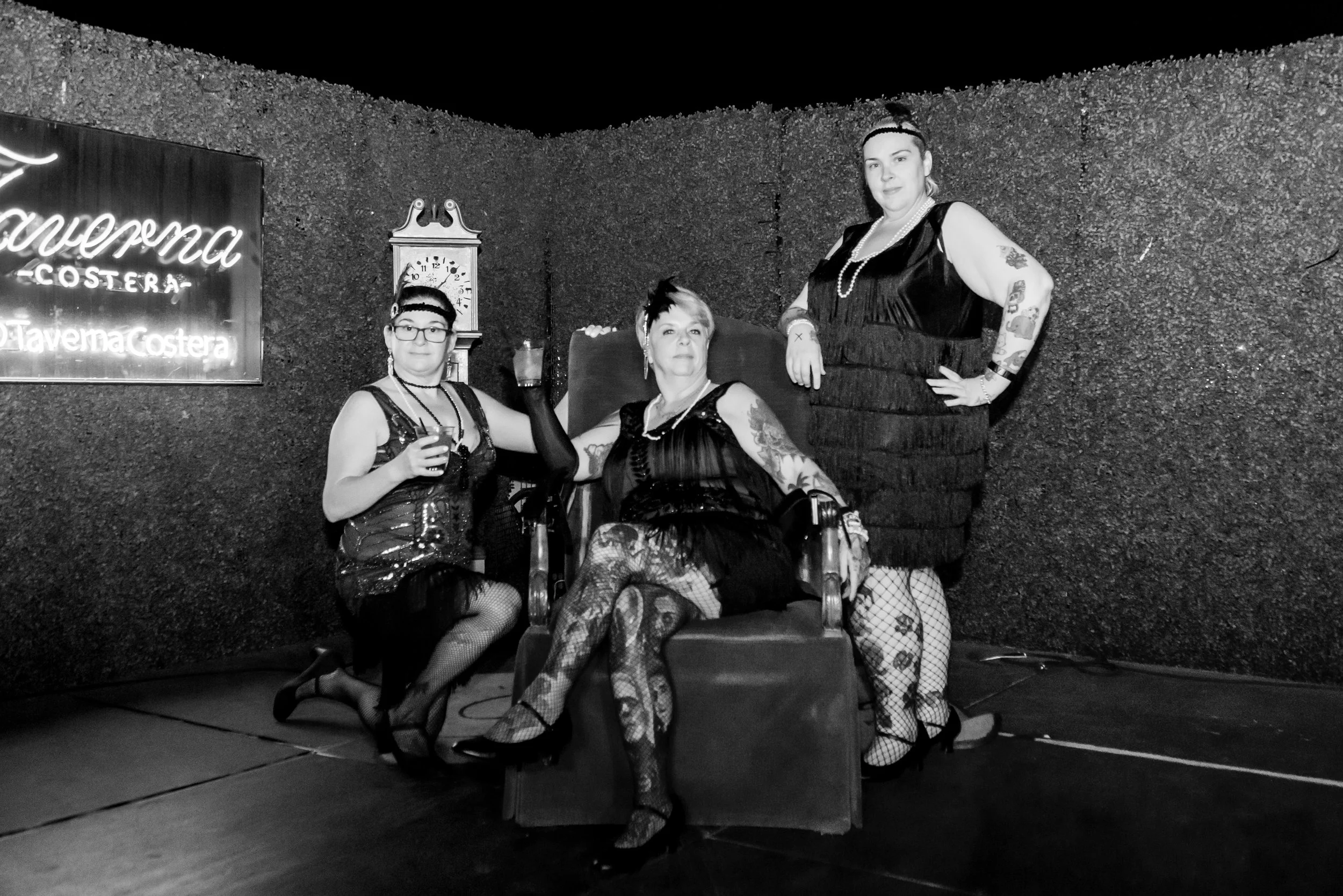 Three women in vintage style clothing and fishnet stockings posing together indoors, with a neon sign and a wall clock in the background.