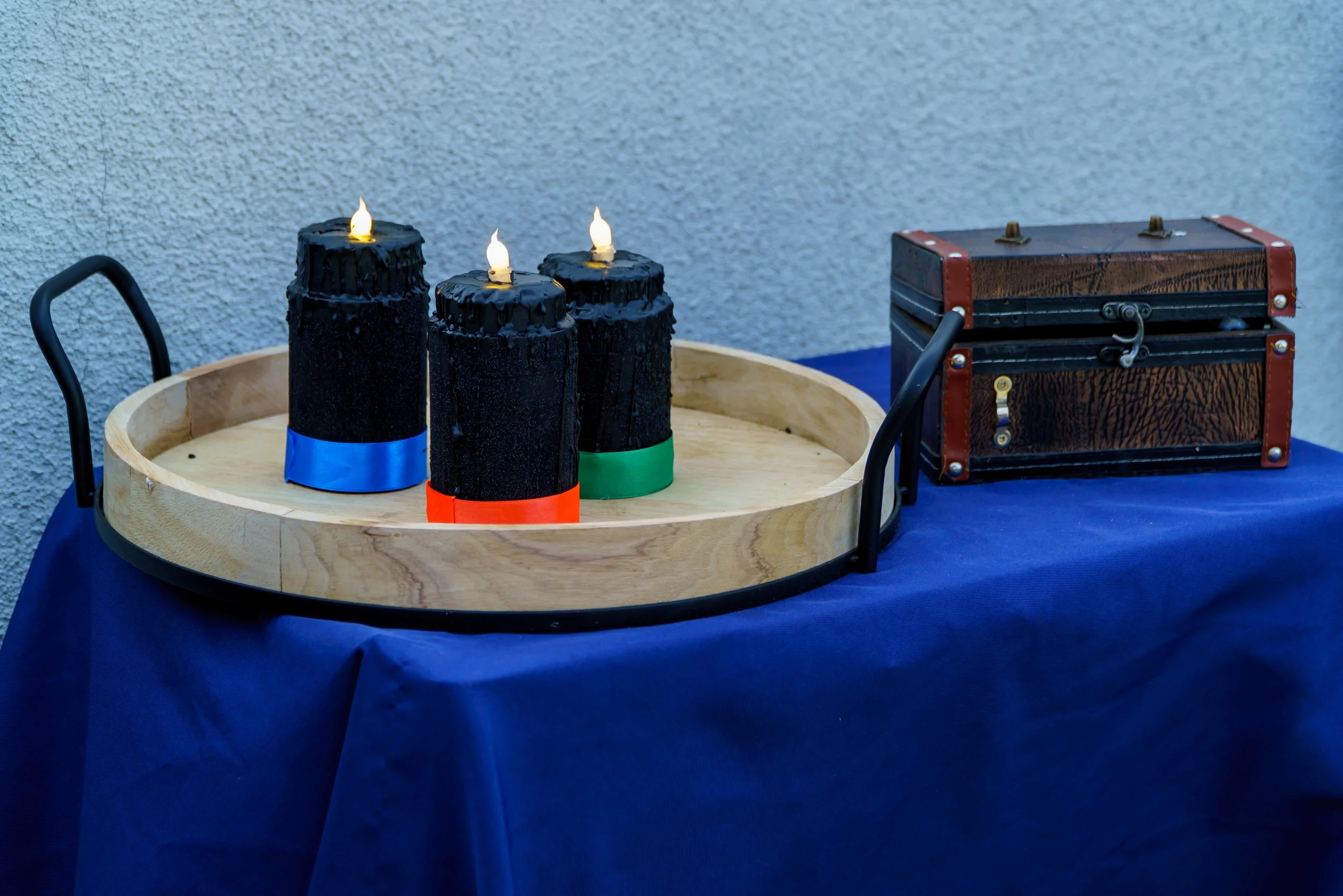 Three black birthday candles with lit flames on a wooden table, decorated with blue, red, and green tape at their bases, next to a small wooden treasure chest on a blue tablecloth against a textured white wall.
