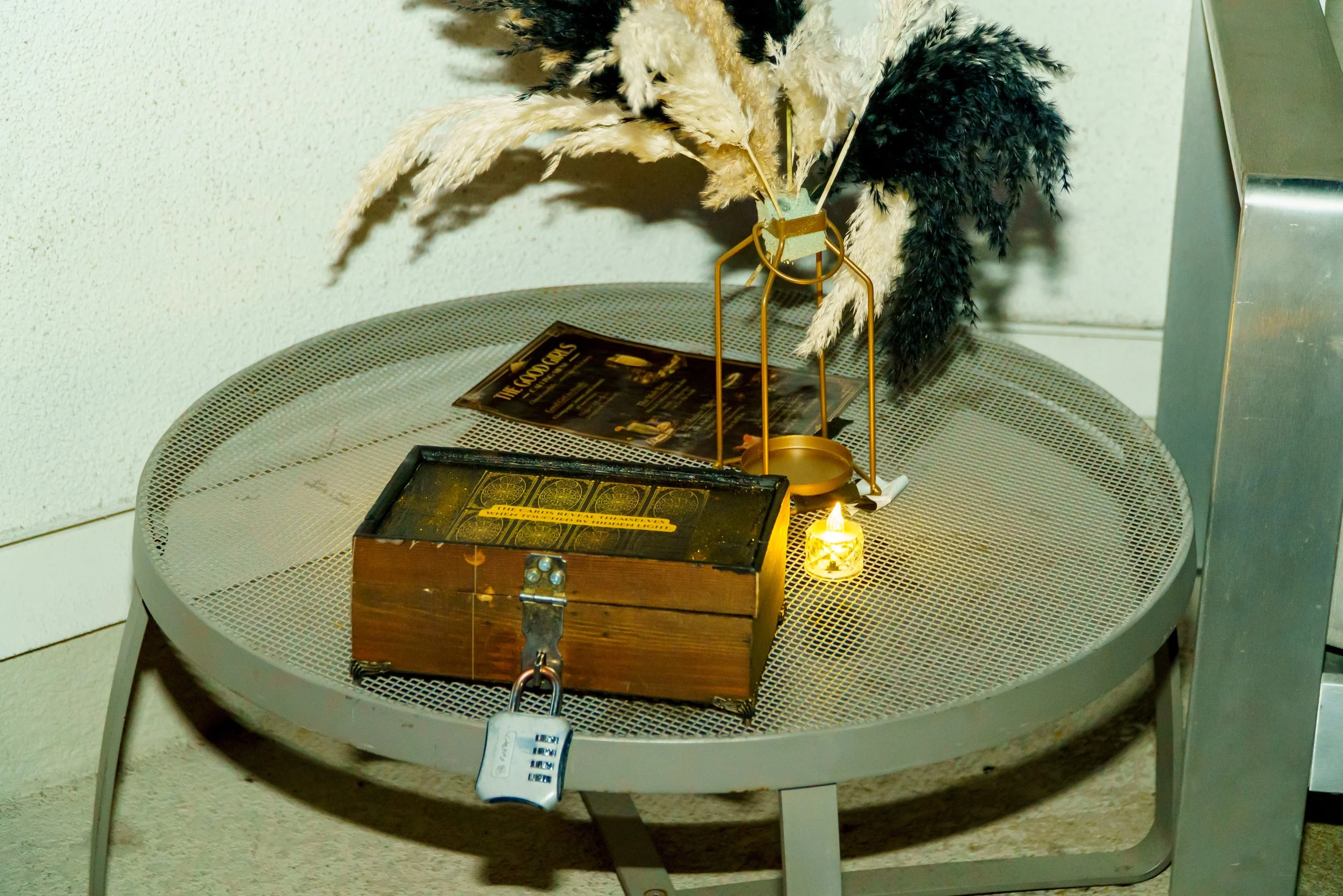 A round metal table with a decorative feather arrangement in a small glass container, a wooden box car with a padlock, a tray with tarot cards, and a small lit candle, all set against a plain wall.