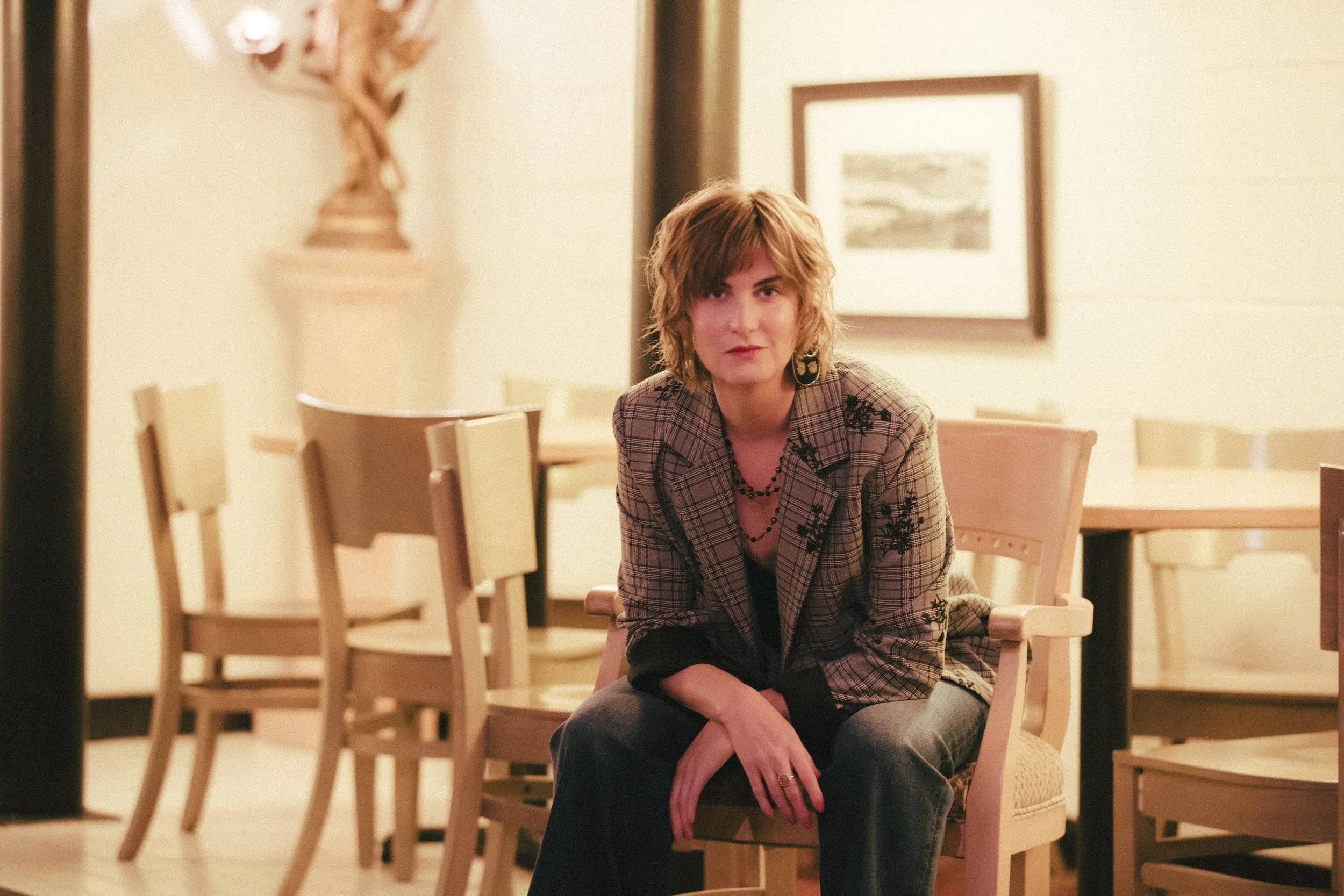 A woman with short, wavy hair, wearing a plaid blazer and black earrings, is seated in a room with beige walls and framed artwork. She is looking at the camera with a slight smile, sitting with one arm resting on her knee.