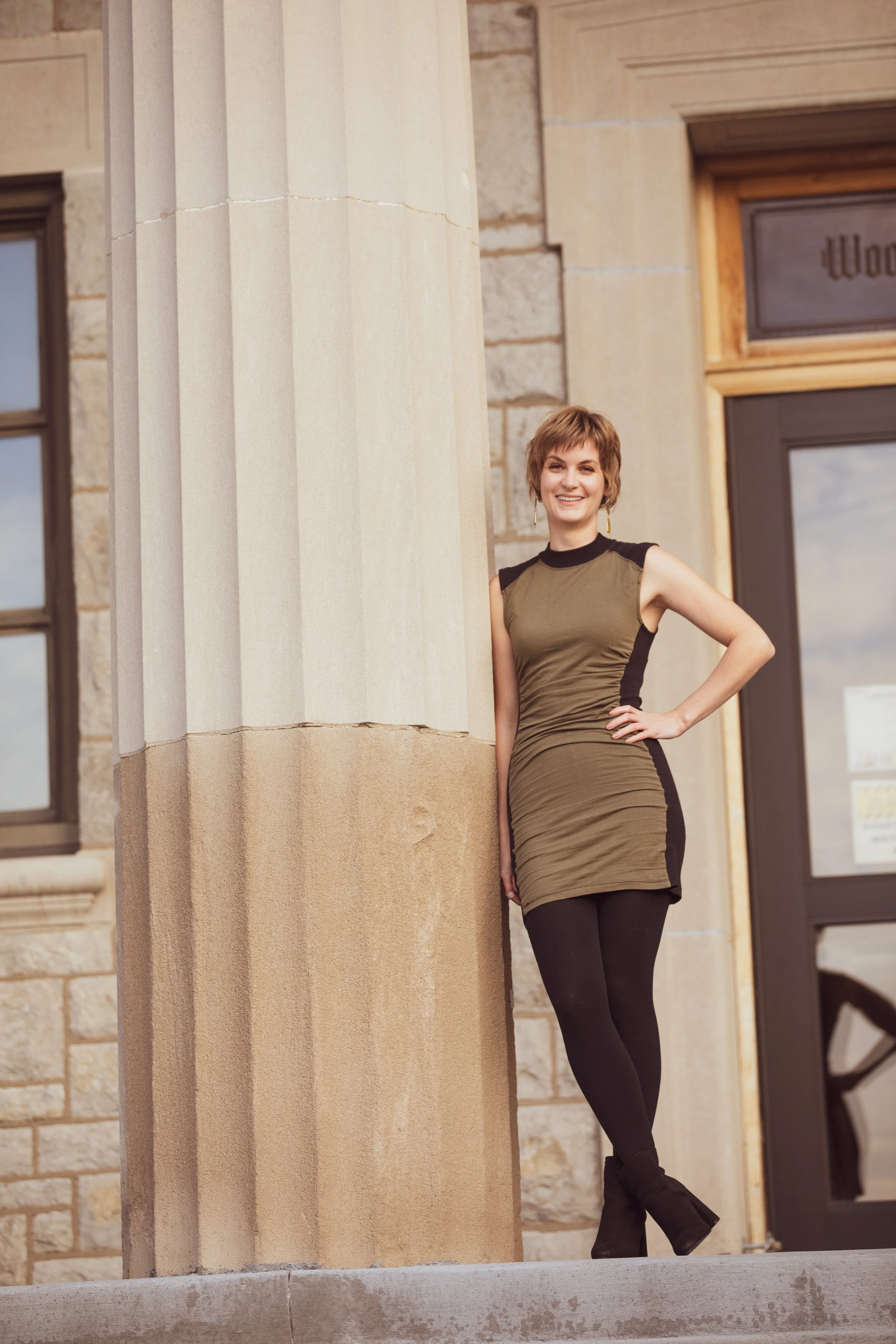 A woman with short hair, wearing a ruffled olive green dress with black accents, black leggings, and black boots, standing against the stone facade of a building with large windows and a door, smiling at the camera.