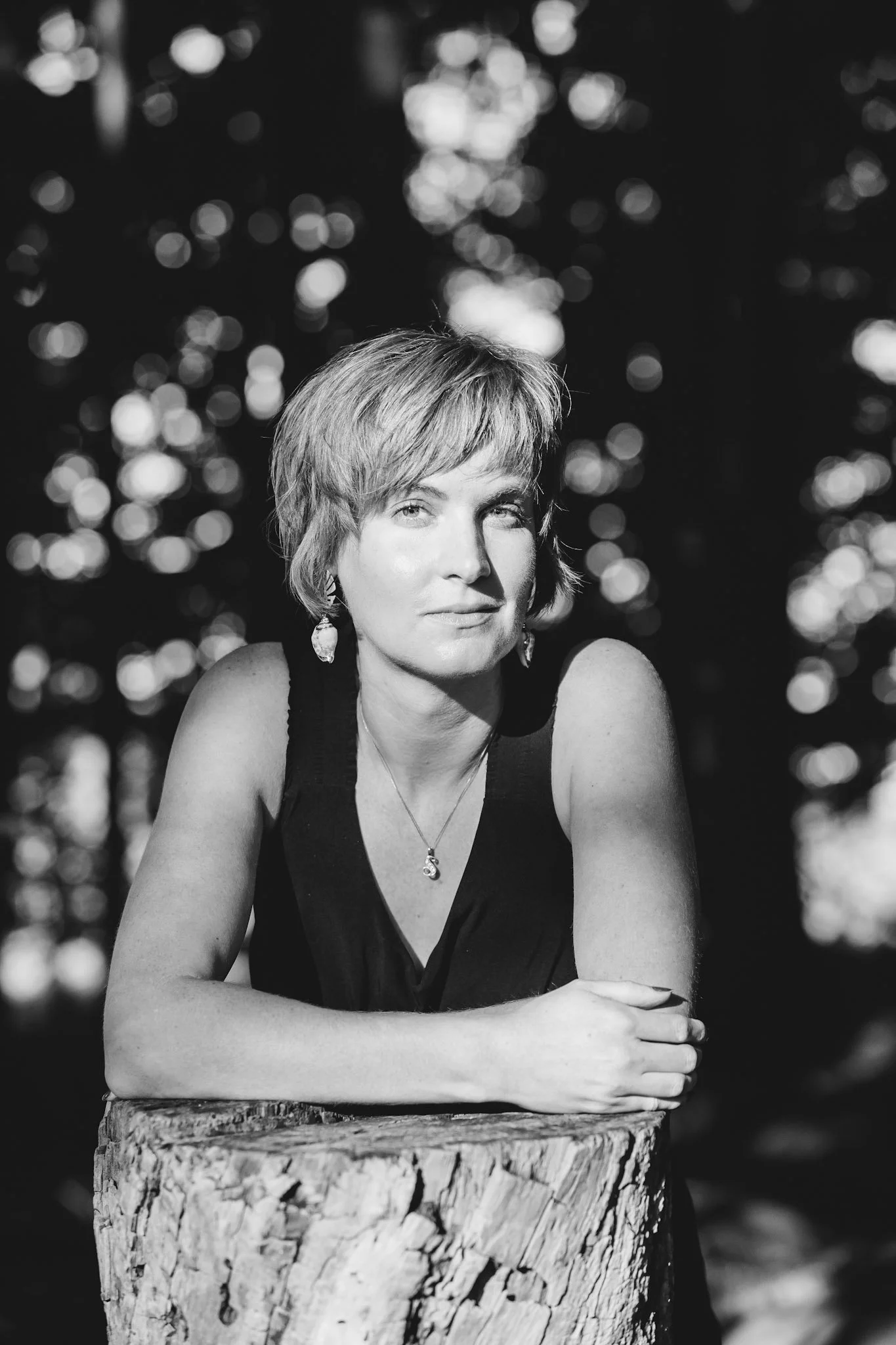 A black and white portrait of a woman with short hair, sitting behind a tree stump, with a blurred background of light reflections on water.