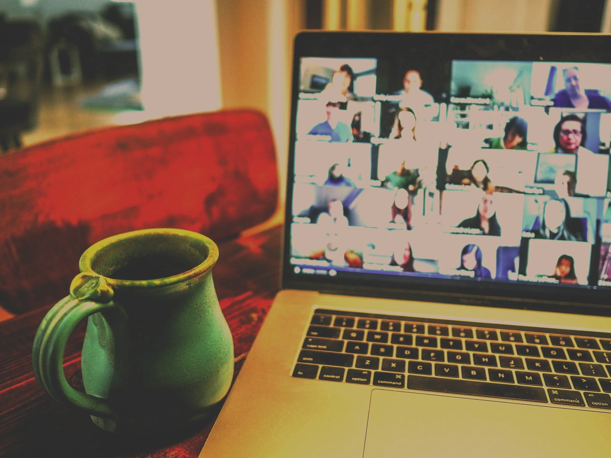 A photo of a coffee mug filled with coffee on a wooden table, in front of a laptop displaying a video call with multiple participants. The background is blurred.