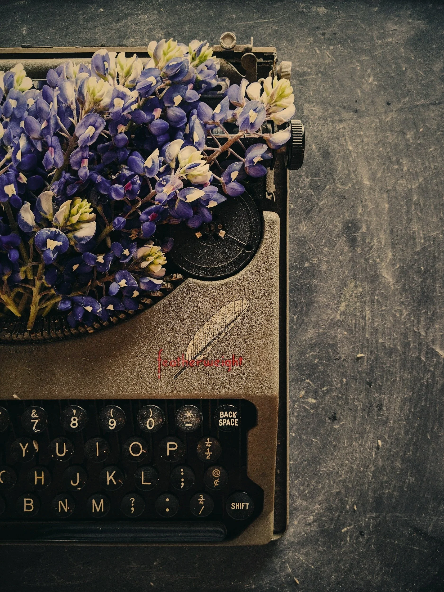 A vintage typewriter with purple and white flowers on top, placed on a dark surface.