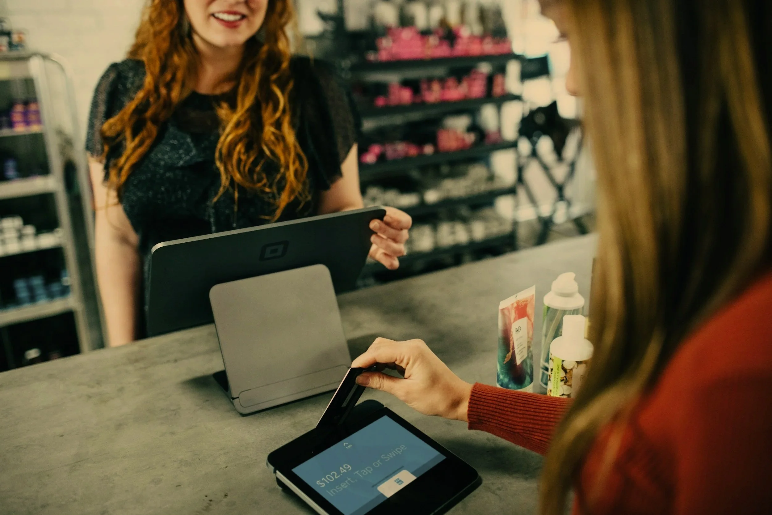 A cashier in a grocery store is scanning items at the checkout counter, while a customer with blonde hair in an orange sweater is paying with a card, and another cashier is smiling and talking to a customer.