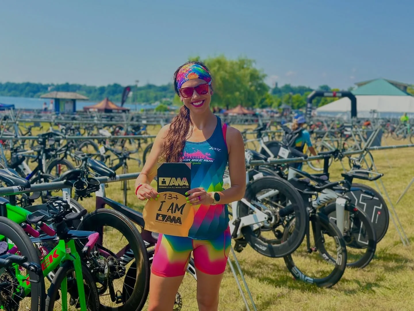 Female athlete holding a race bib and a plaque, standing in front of a row of bicycles on a grassy field during a sports event.