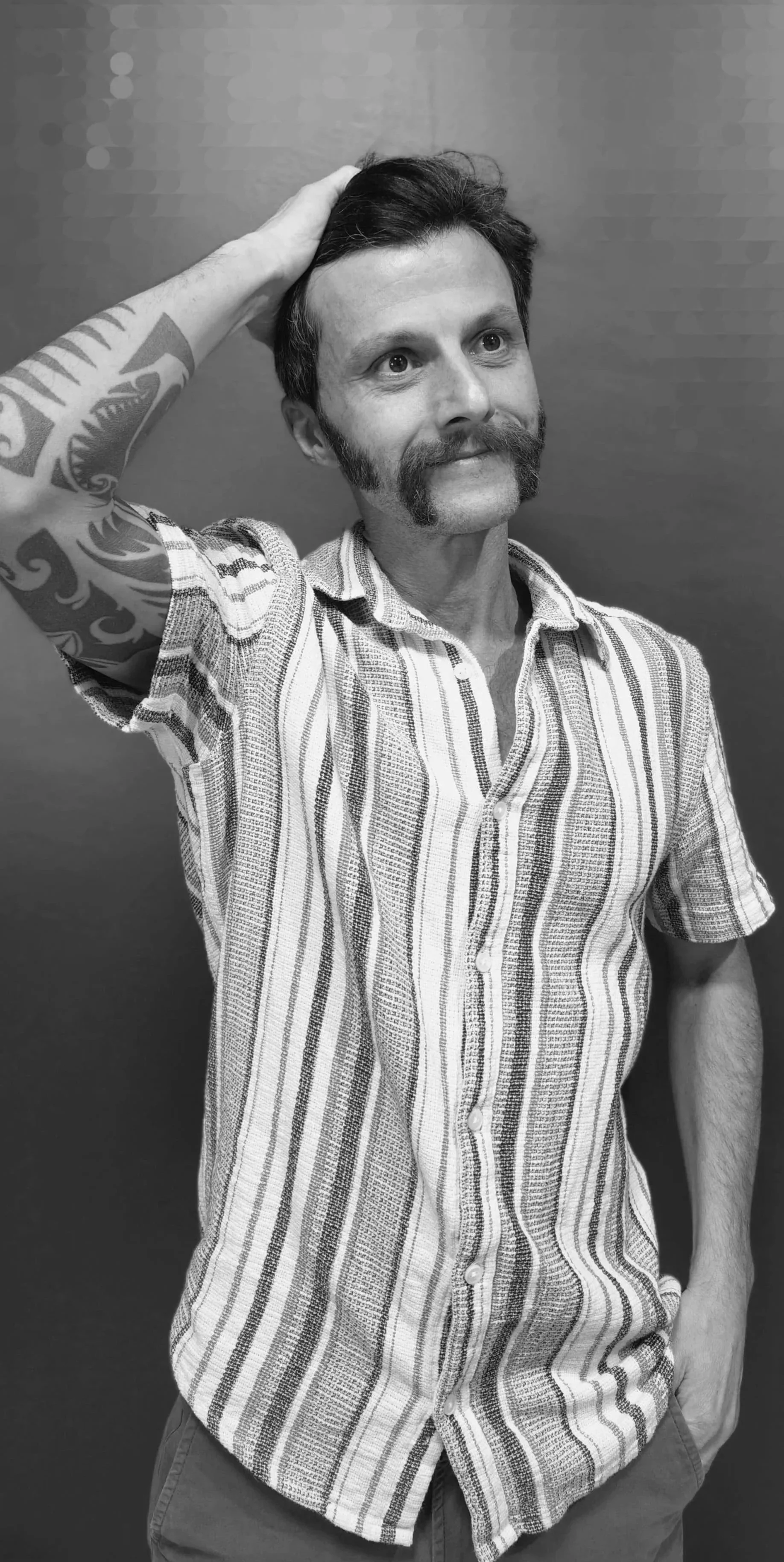 A man with beautiful side burns and a style moustache, wearing a striped short-sleeve shirt, standing against a plain wall. He is touching his head with his right hand, which has an amazing tattoo sleeve.