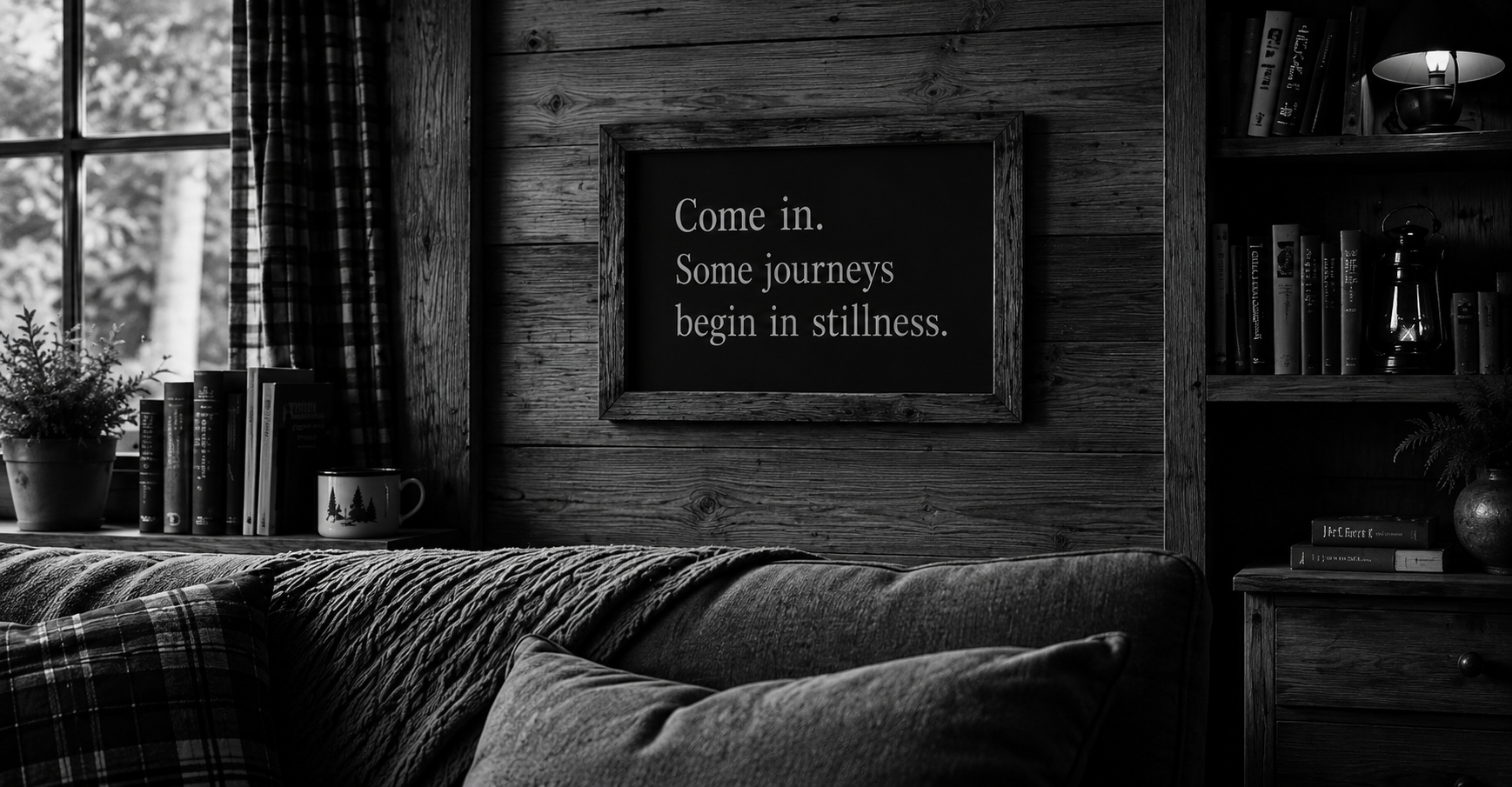 A cosy wooden interior room with a window and a bookshelf, featuring a framed quote on the wall that reads, "Come in. Some journeys begin in stillness."