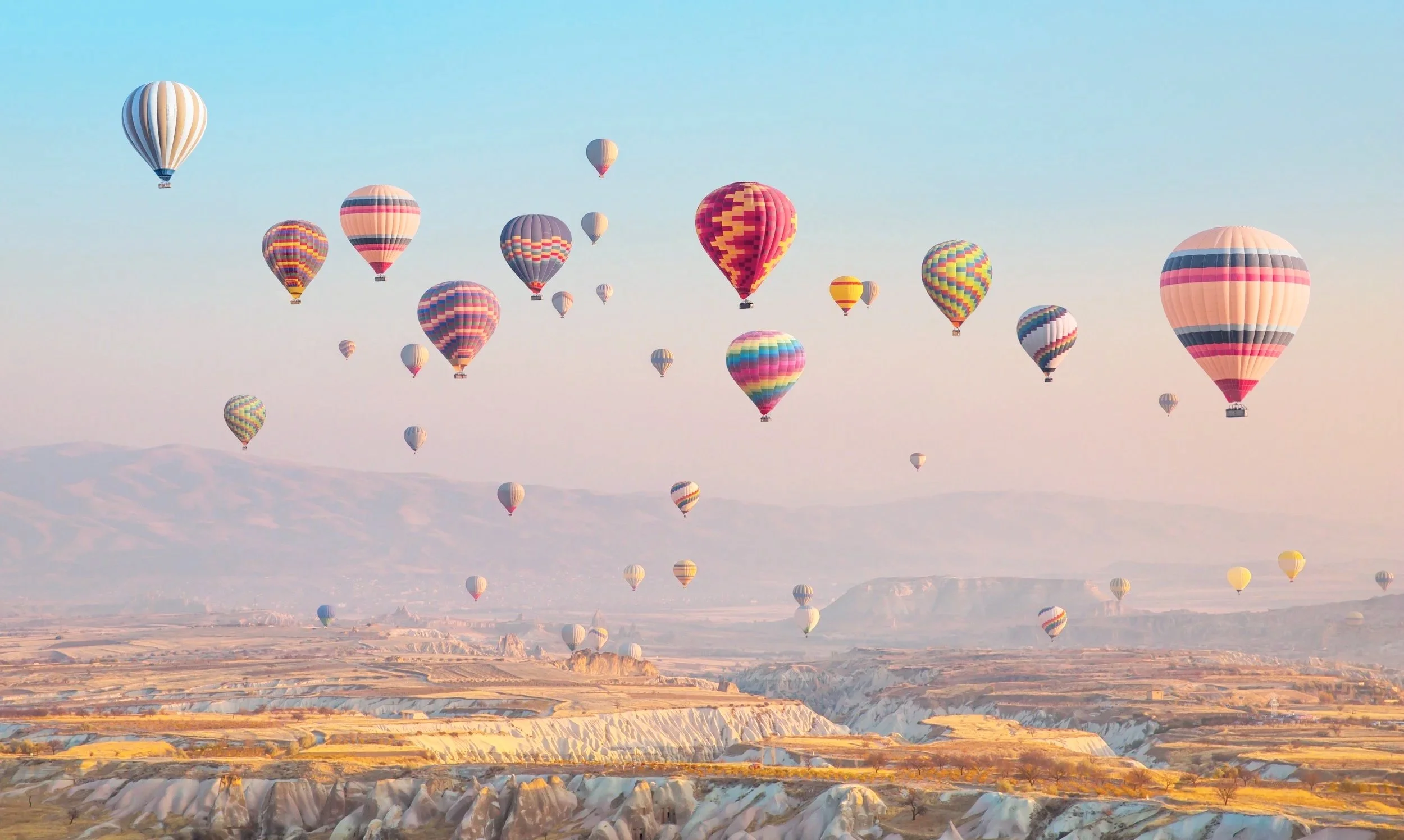 Multiple colorful hot air balloons floating in the sky over a desert landscape with rock formations and mountains in the background during daylight.