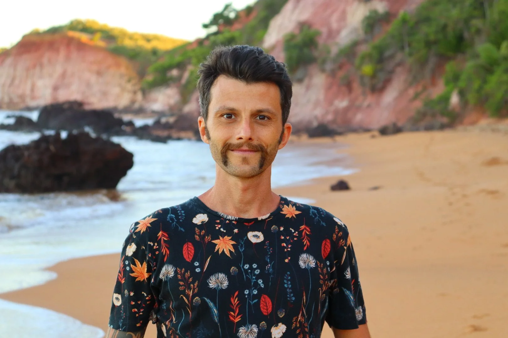 A man with dark hair, a pretty unique elbow tattoo, a styled horseshoe's moustache and beautiful sideburns stands on a sandy beach with cliffs and greenery in the background.