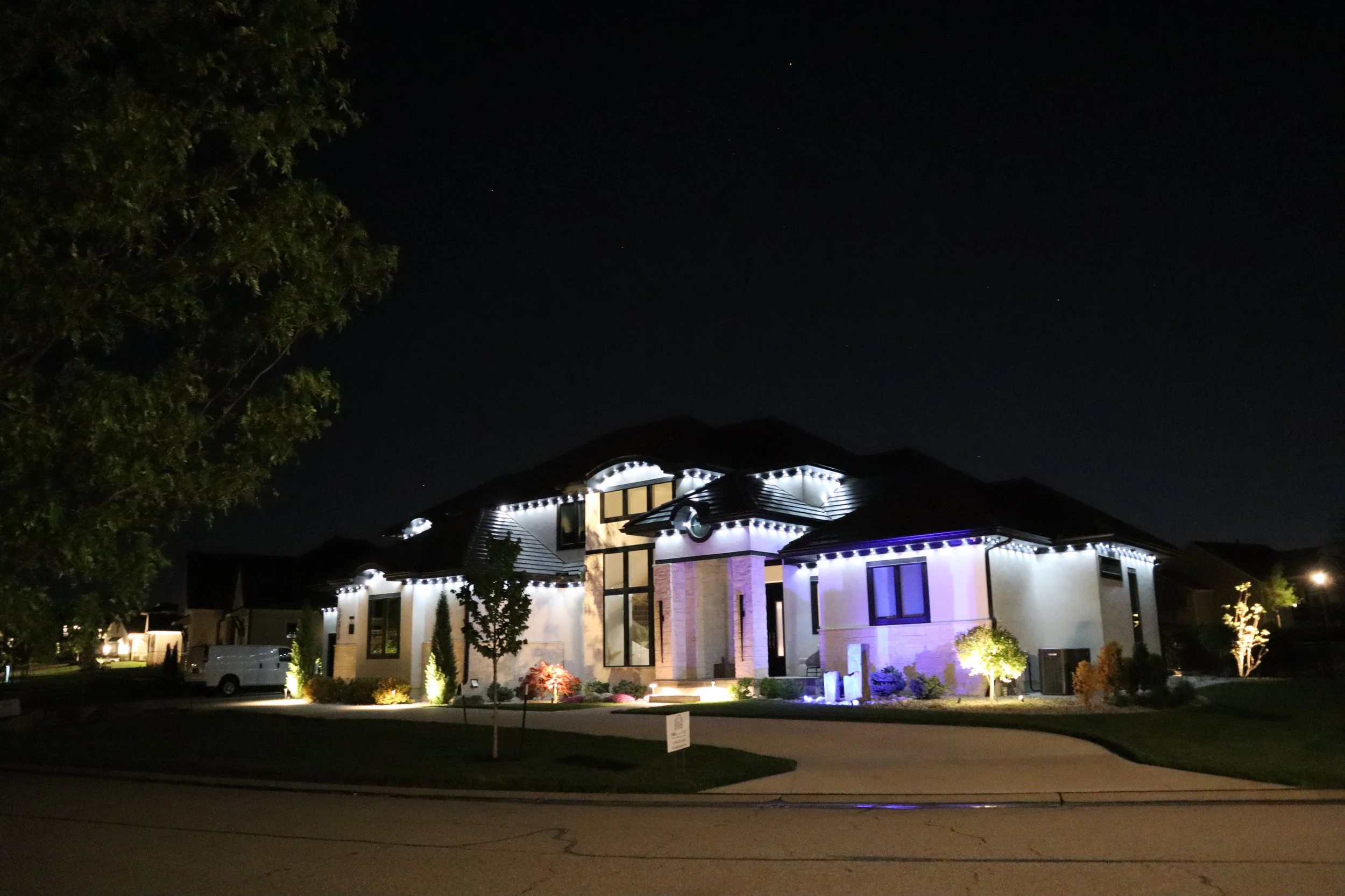 Night view of a modern house with white exterior illuminated by exterior lights, large windows, and decorative lighting on the roof and landscape, with a tree on the left and a dark sky with stars.