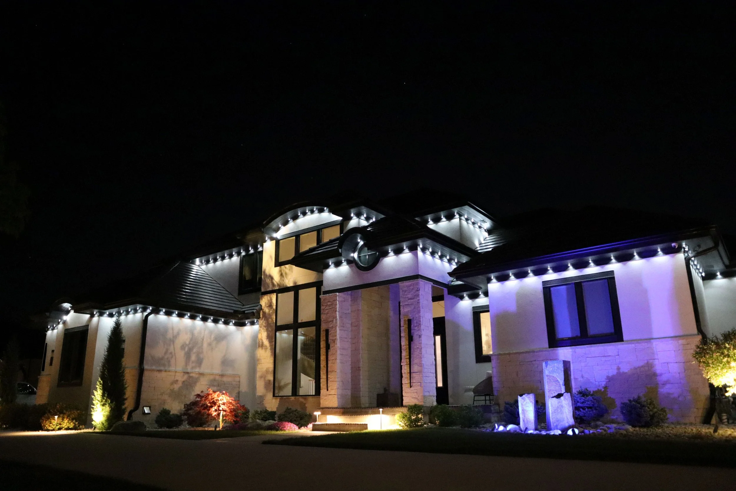 Night view of a modern house illuminated with exterior lights, showcasing architectural details, with a well-maintained garden in the foreground and a dark sky above.