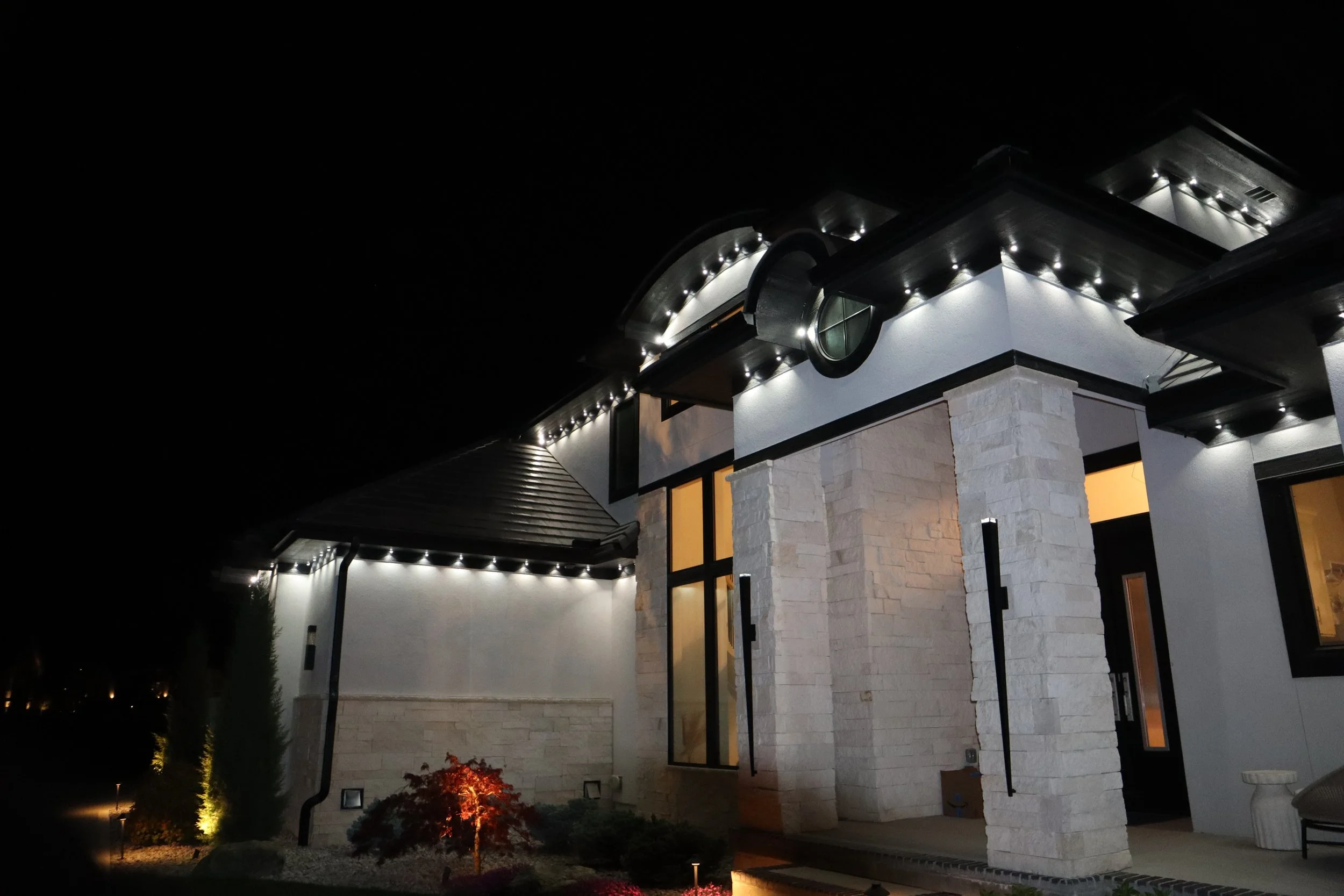Night view of a modern house with exterior lights, stone and stucco walls, and large windows, illuminated against a dark sky.