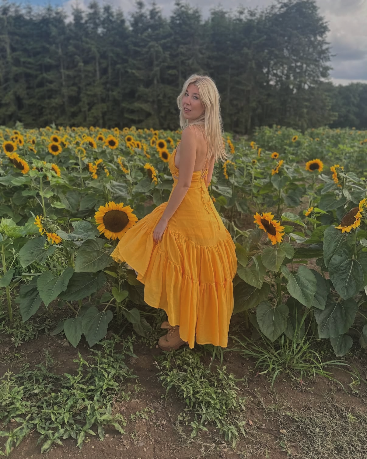 A woman in a flowing yellow dress standing in a sunflower field with green foliage and trees in the background.