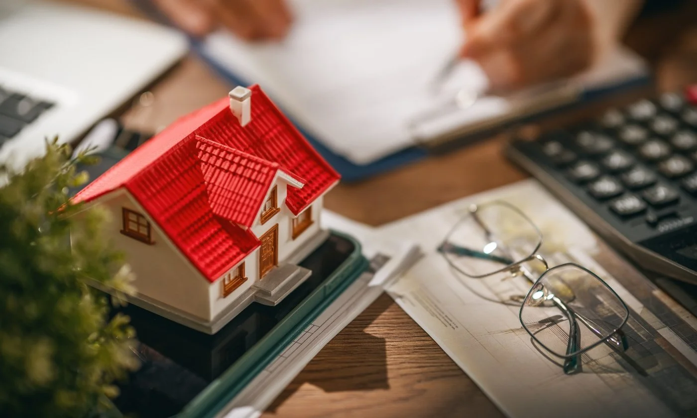 Model house with red roof on an office desk, surrounded by paperwork, eyeglasses, calculator, and pen.