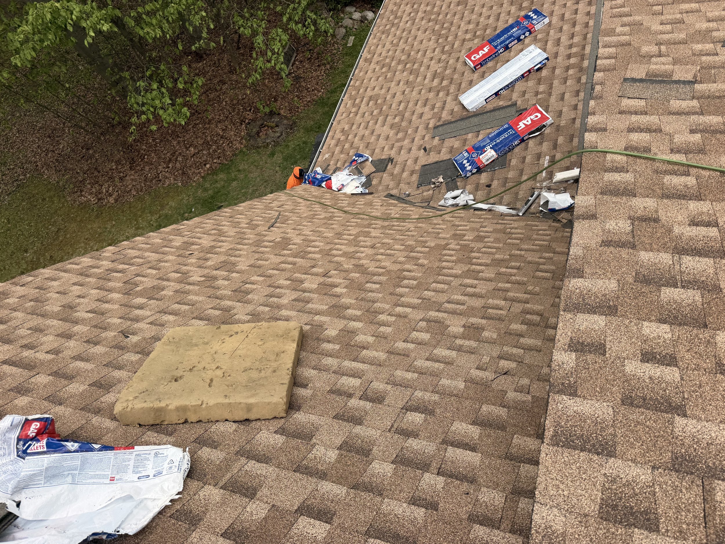 A rooftop with shingles, debris, and torn packaging materials scattered across the surface. On the left, a tree with green leaves is visible, and on the right, a beige stone or concrete block rests on the shingles.