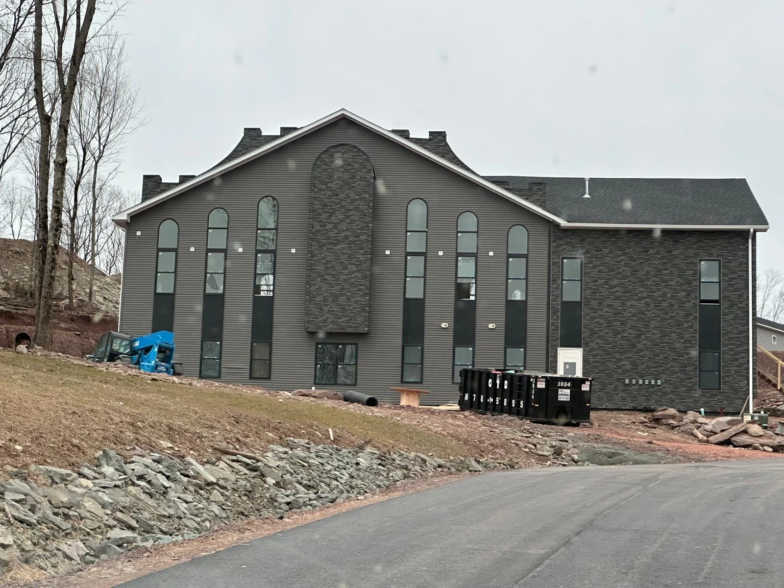 A large gray house under construction with a black dumpster in front. The house has tall, narrow windows and a textured brick and siding exterior. The surrounding area is rocky and muddy with construction materials and a small blue lift near the house.