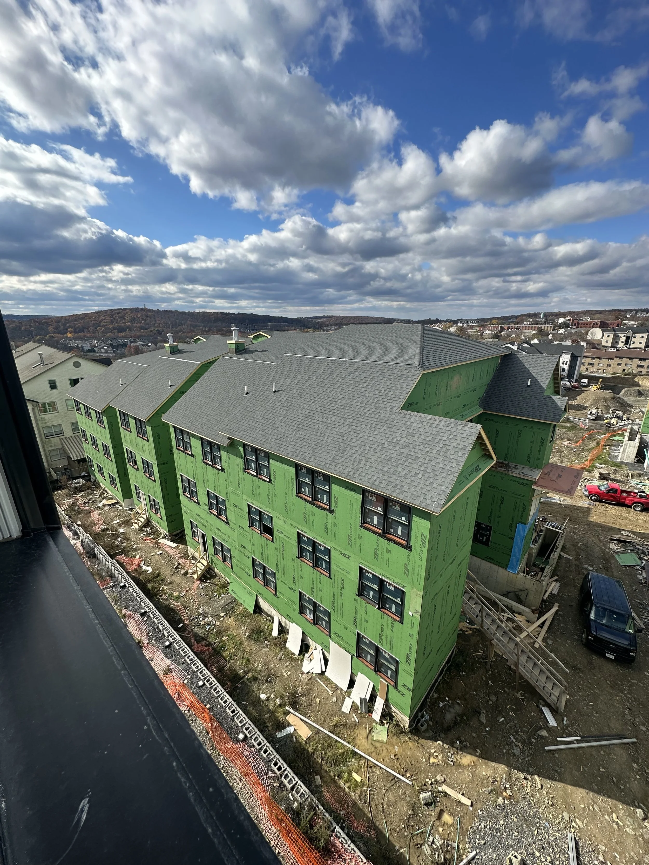 Construction site with a partially built multi-story green building, surrounded by construction materials, equipment, and safety barriers, under cloudy skies.