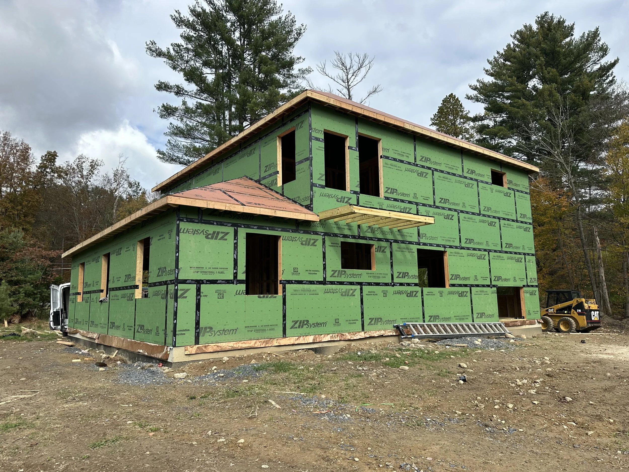 A two-story house under construction with green ZIP System sheathing, surrounded by trees with autumn foliage, construction equipment, and a cloudy sky.