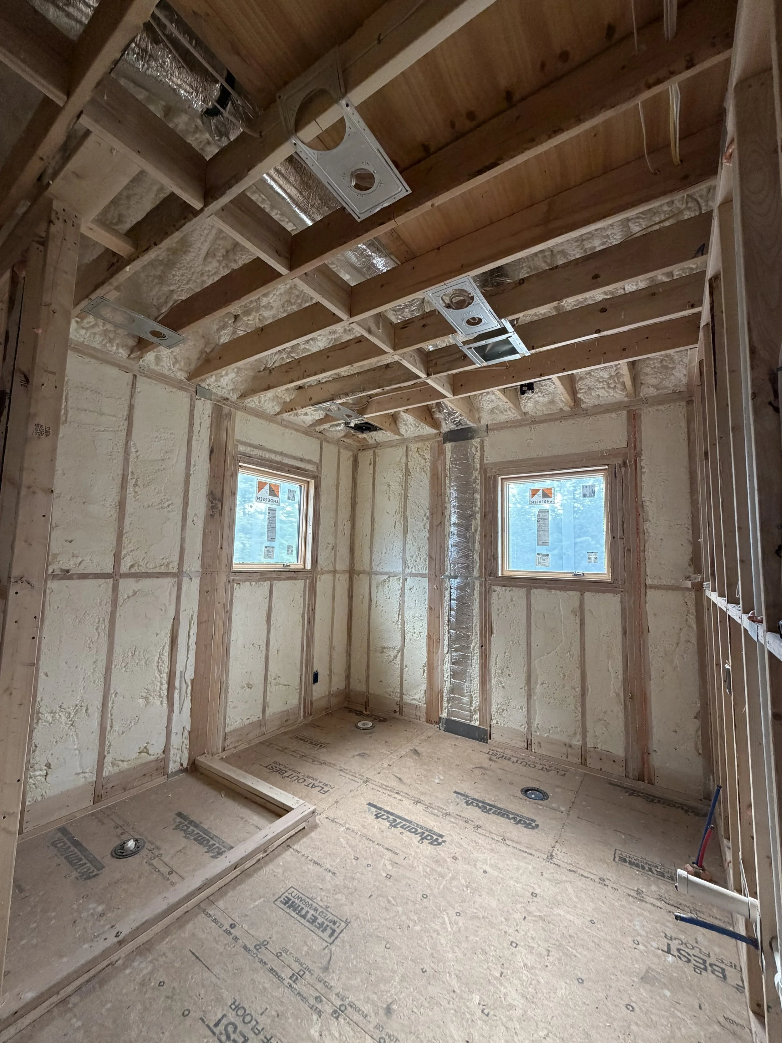 Interior of a room under construction with exposed wooden framing, insulation, and two windows.