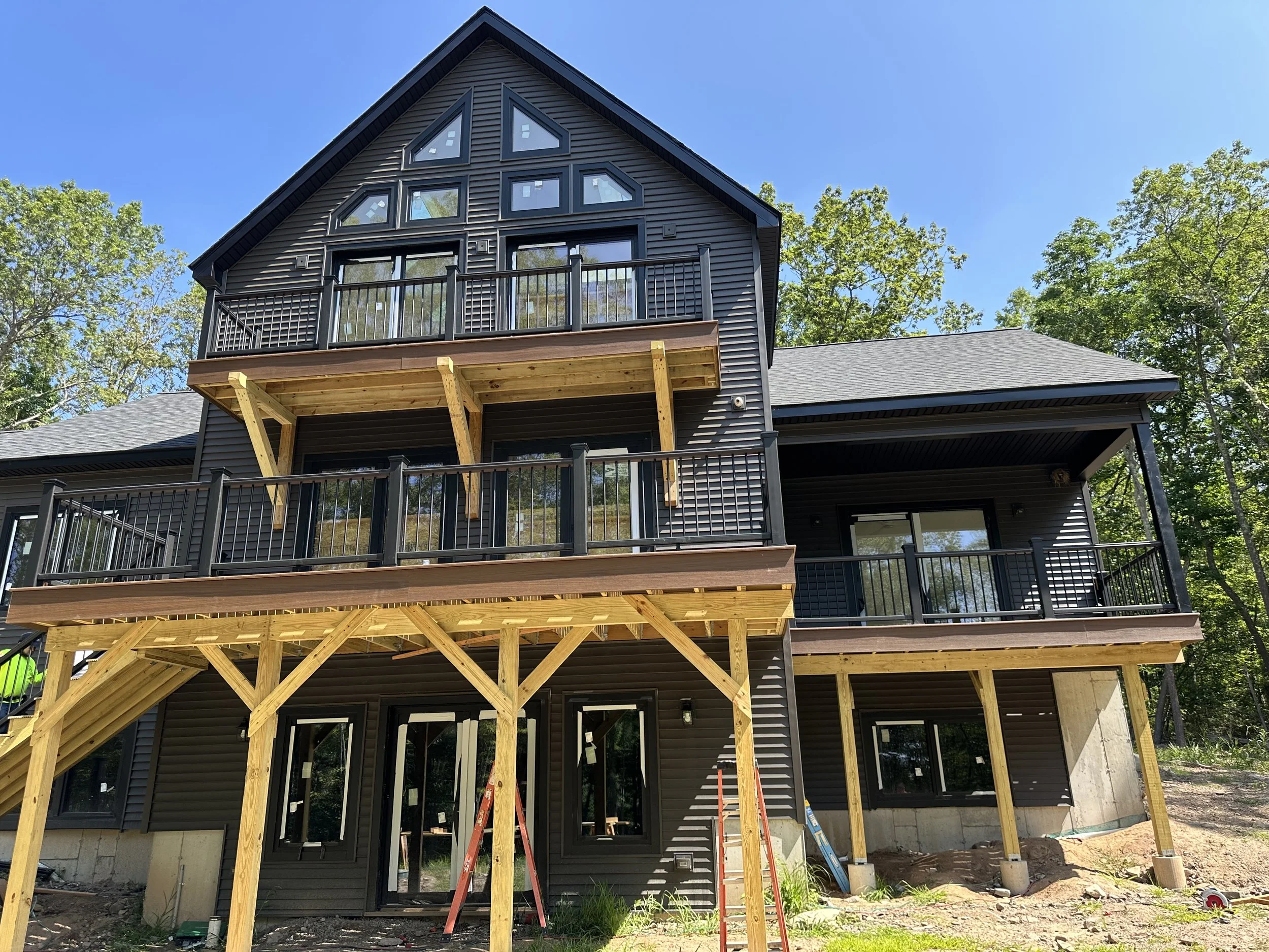 A house under construction with a black exterior, multiple balconies, and large windows, surrounded by green trees against a blue sky.