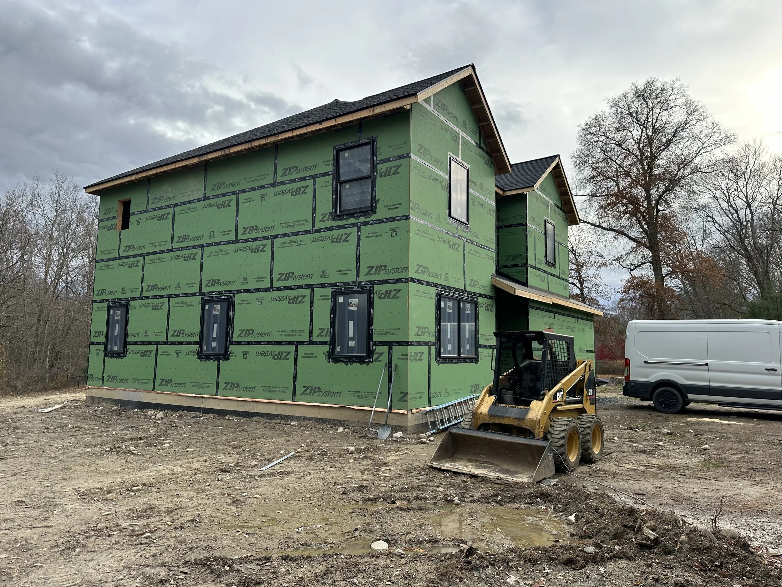 A house under construction with green sheathing, several windows installed, and construction equipment on the site.