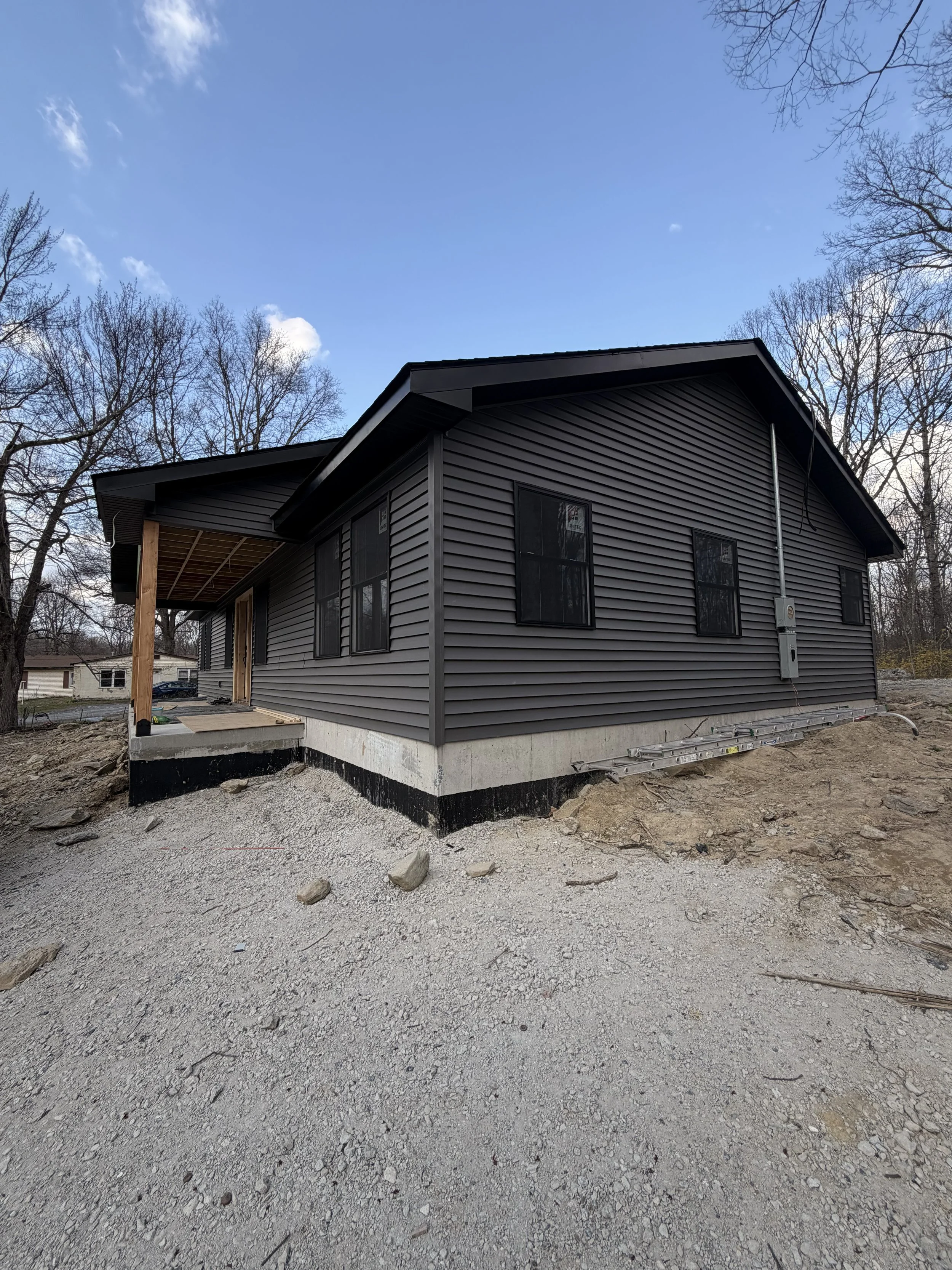 Side view of a house under construction, with dark gray vinyl siding, black window frames, and an incomplete porch area, set on a gravel foundation with dirt ground in a wooded area.