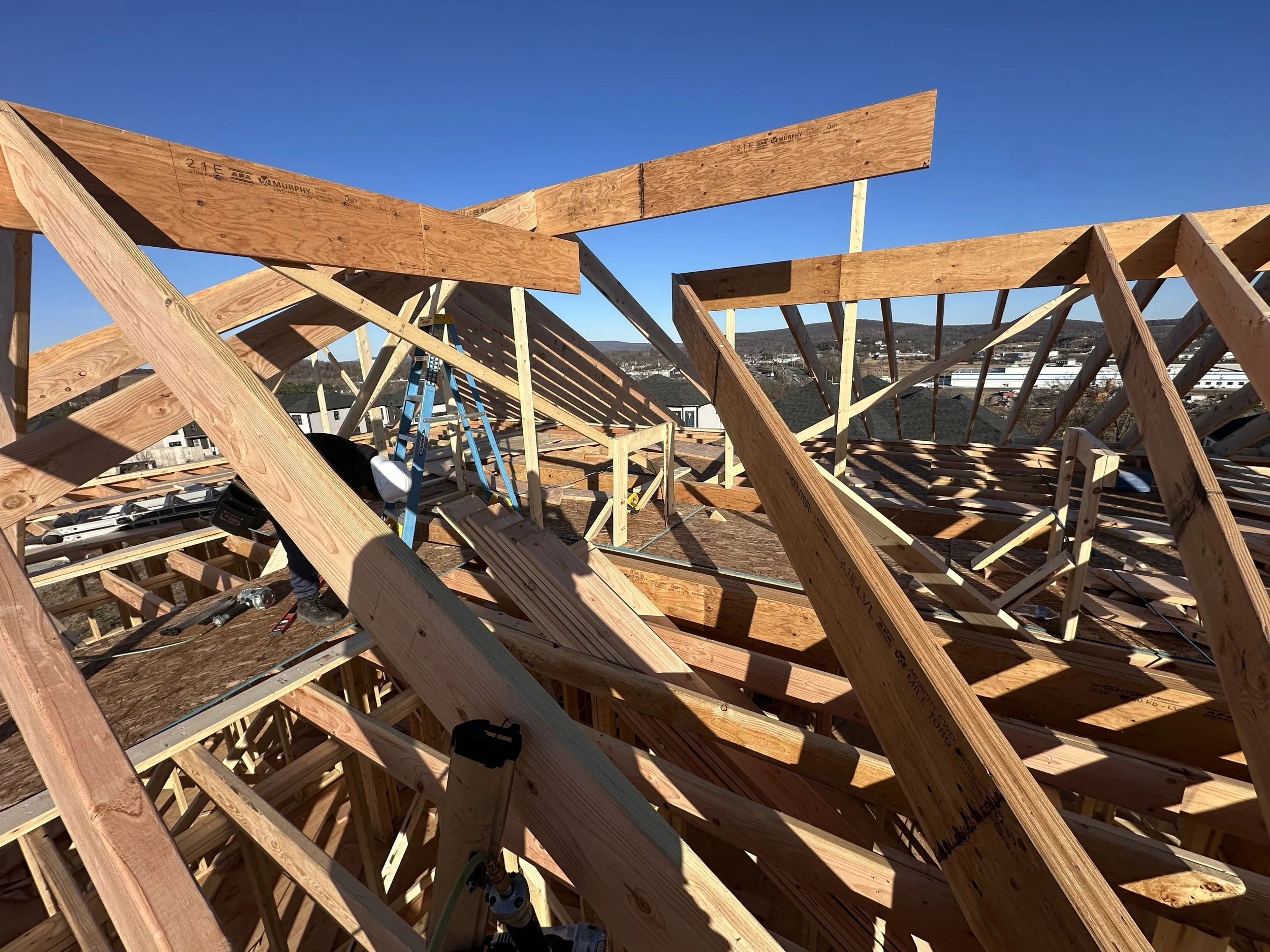 Construction site with wooden framing for a building, including beams and a ladder, under a clear blue sky with a background of distant hills and buildings.