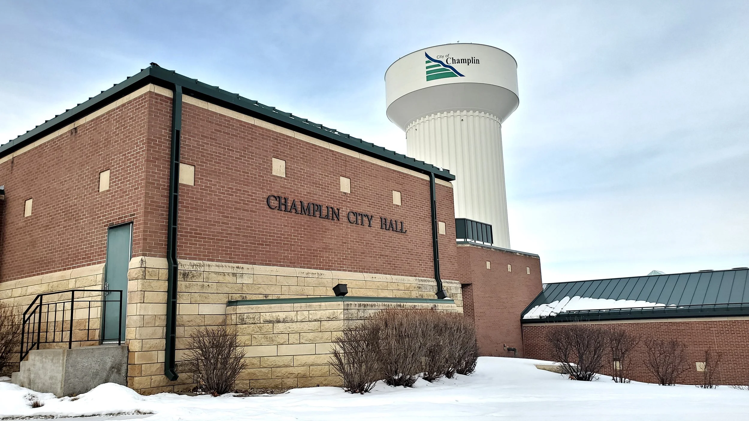 Image of Champlin City Hall building with a large water tower in the background, and snow on the ground.