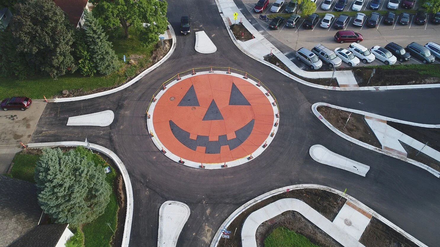 A bird's-eye view of a roundabout with a large Halloween pumpkin face painted in orange and black at the center. The roundabout is surrounded by parking lots and green trees.
