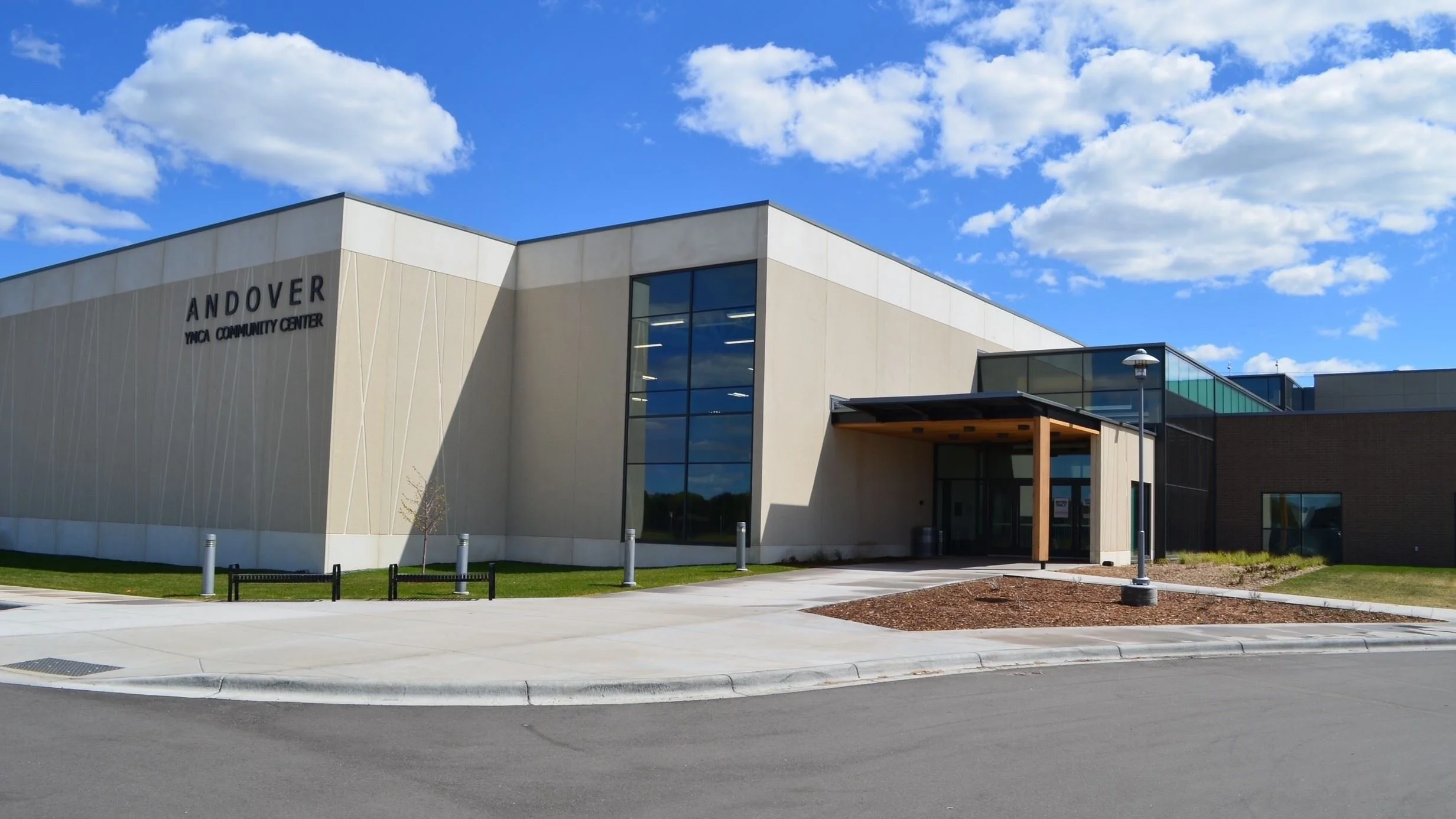 Modern building with large glass windows and beige walls, identified as Andover YMCA Community Center, under a partly cloudy blue sky.
