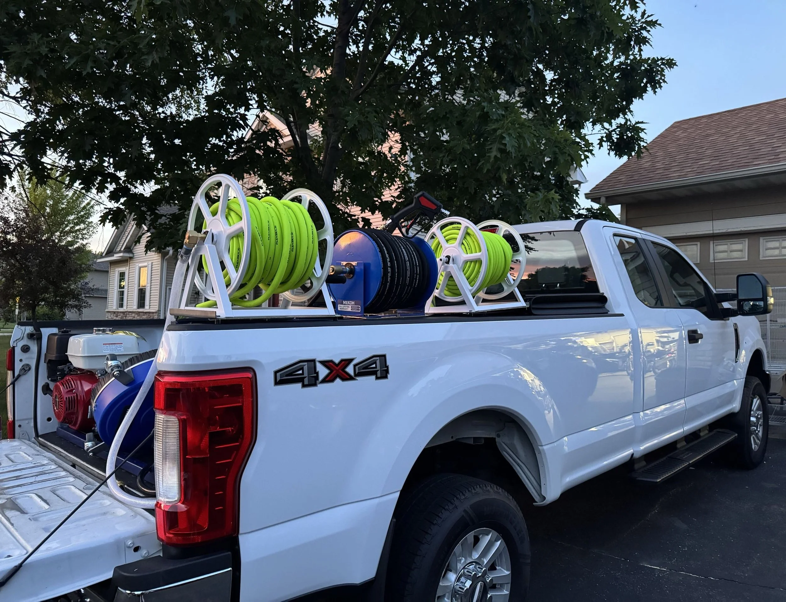 A white pickup truck with a ladder rack carrying hoses and equipment, parked in a residential neighborhood with trees and houses in the background.