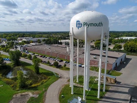Wide aerial view of a water tower with the Plymouth logo, surrounded by industrial buildings, parking lots, and green trees on a partly cloudy day.