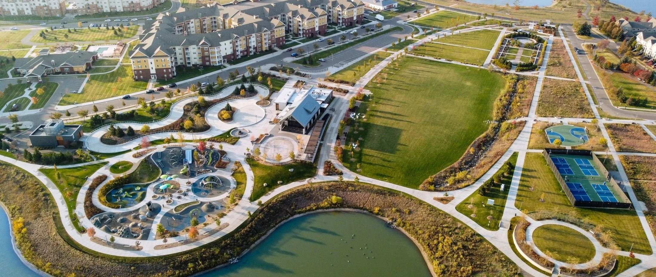 Aerial view of a modern residential complex with landscaped parks, walking paths, water features, playgrounds, tennis courts, and parking lots.